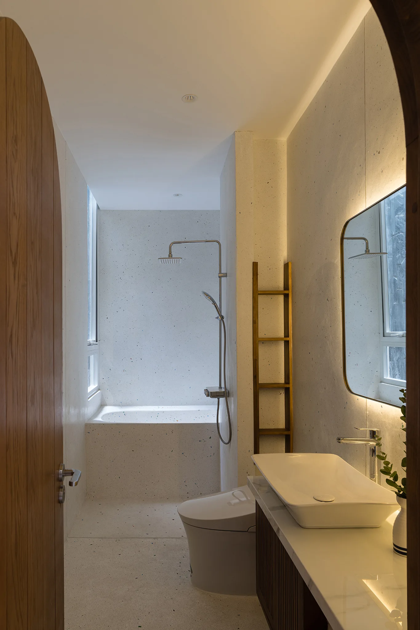 A modern bathroom seen through an arched wooden doorway, featuring speckled terrazzo walls, a built-in tub, and a vanity with a backlit mirror.