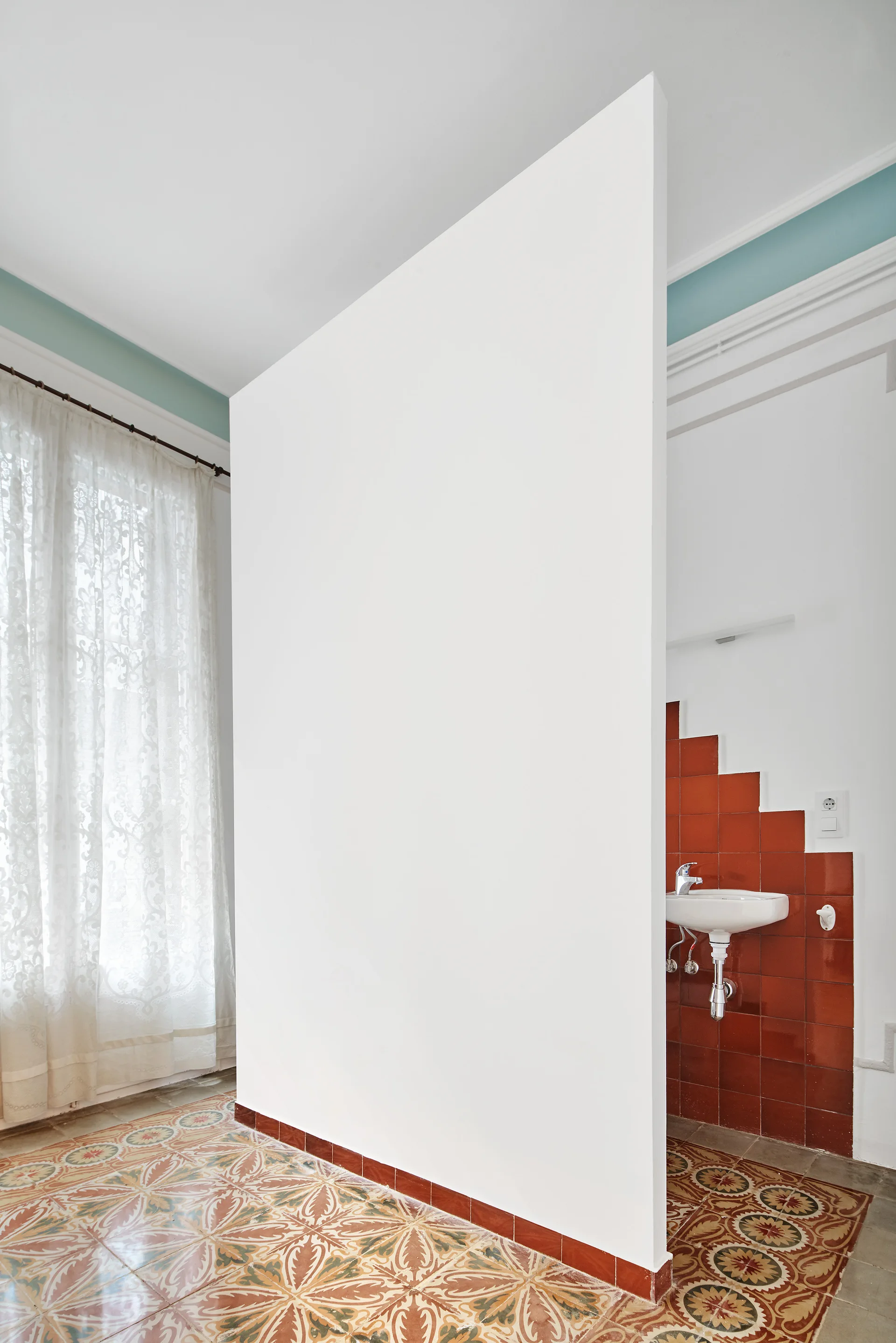 A white partition wall divides a bathroom, separating a lace-curtained window from a wall-mounted sink with red terracotta tile and patterned flooring.