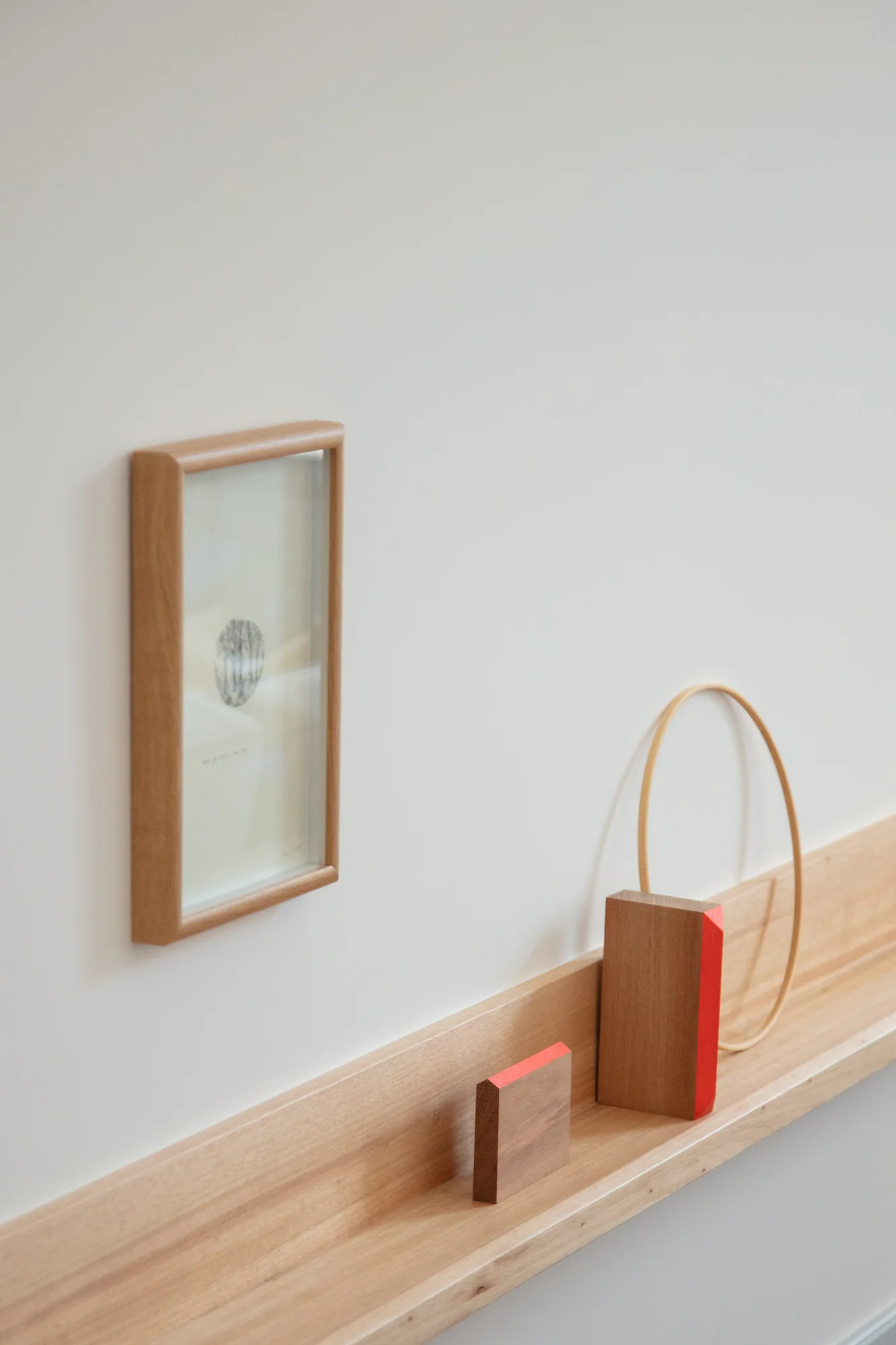 A light wood floating shelf holds geometric wooden blocks and a thin hoop against a white wall next to a small framed picture.