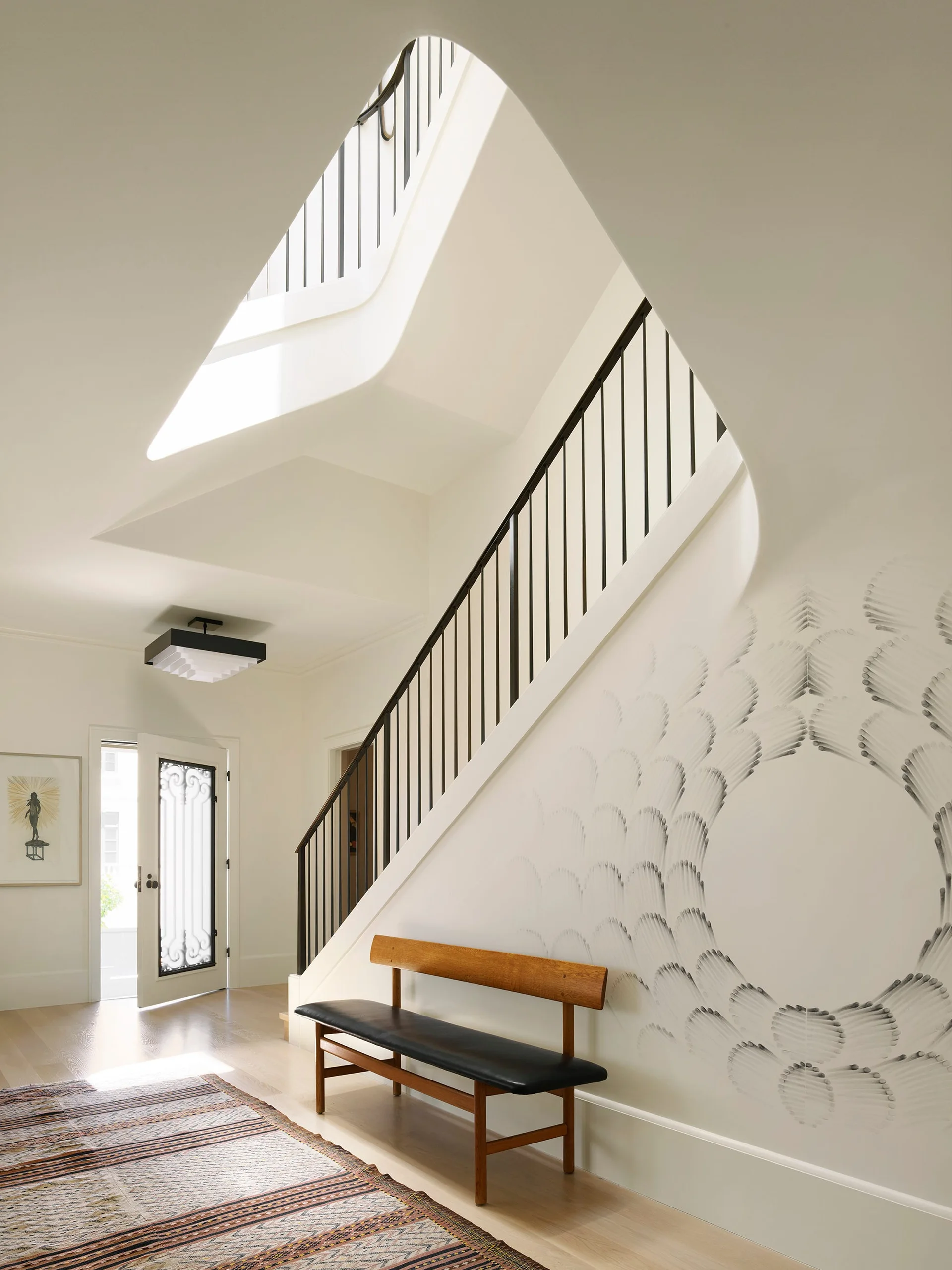 A bright foyer featuring a curved staircase with black railings, an abstract wall mural, a wooden bench, and light wood flooring.