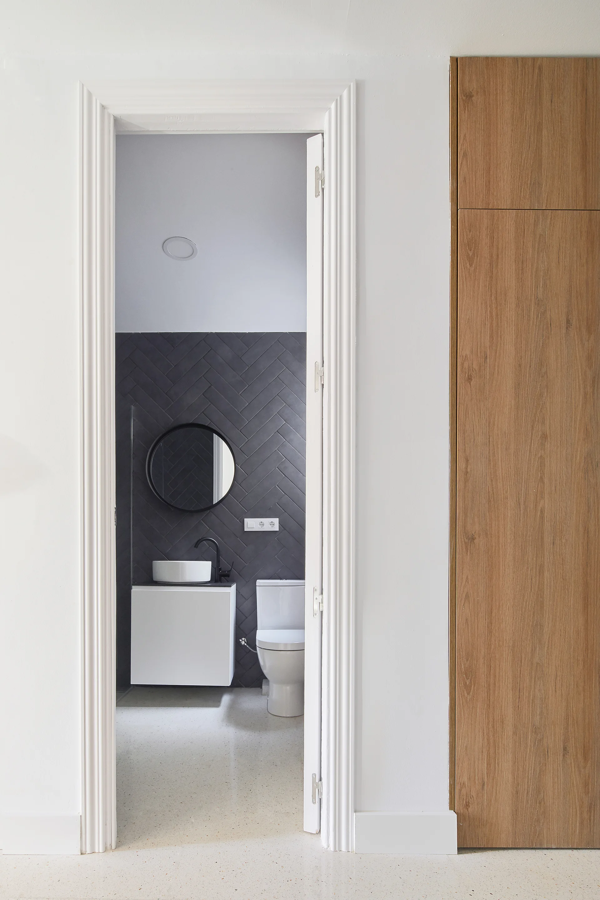 A view through a white doorway reveals a modern bathroom featuring a charcoal herringbone tile wall, a round black mirror, and a white floating vanity.