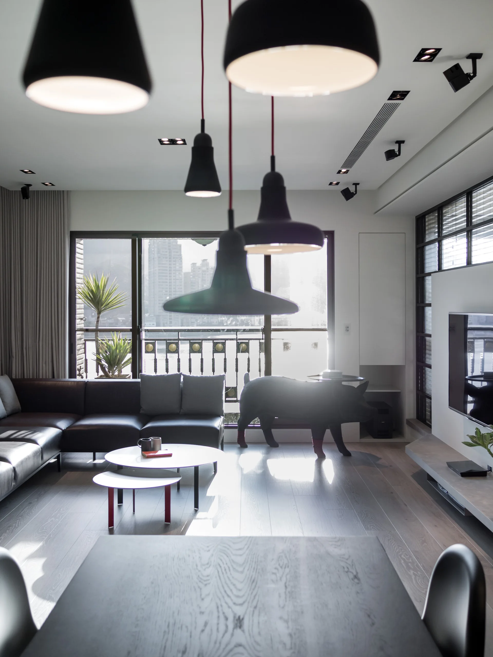 A contemporary living room with a dark leather sectional sofa, matte black pendant lights, and nesting coffee tables in front of a window.