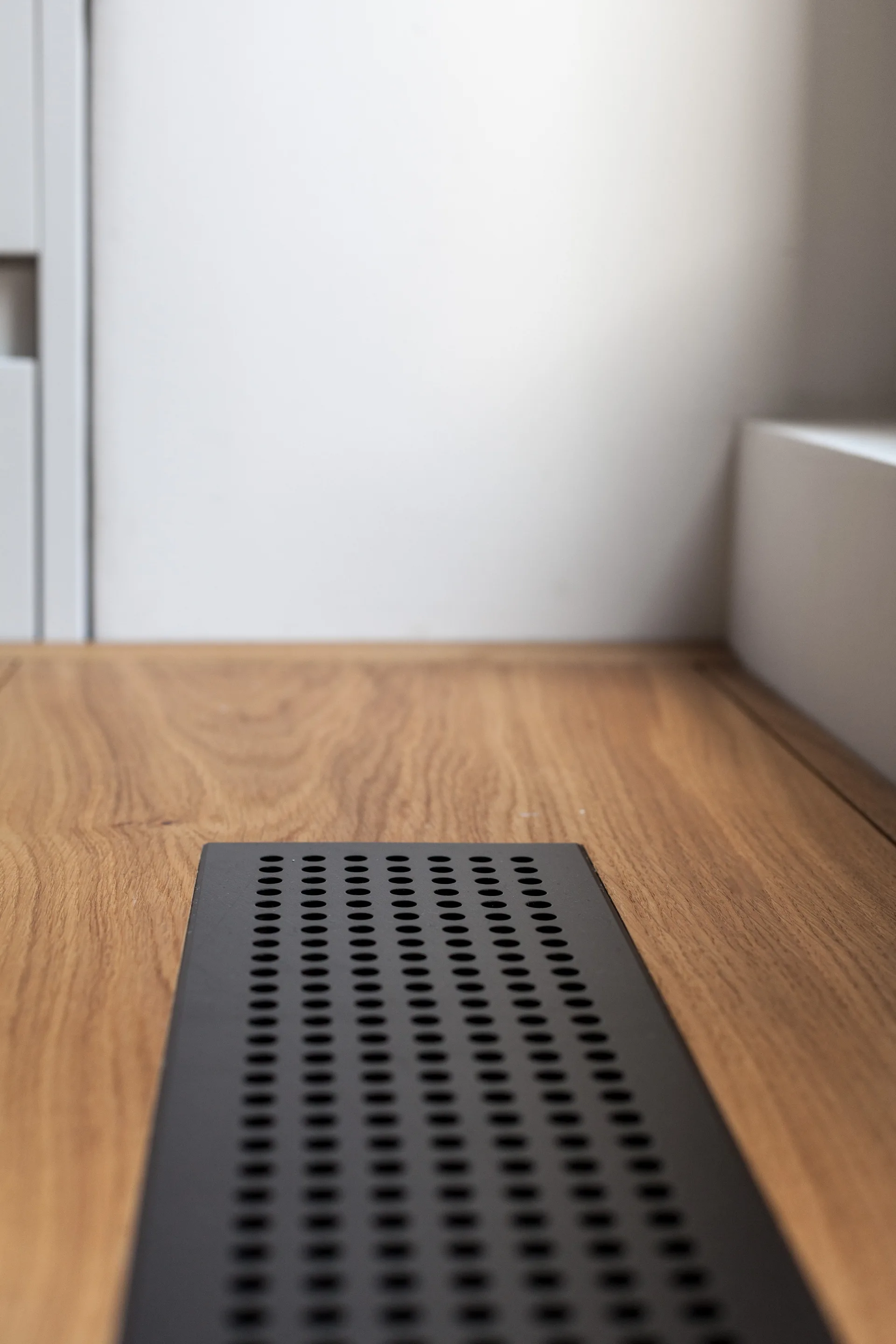 A close-up of a black perforated metal floor register seamlessly embedded in a light wood plank floor beside a white wall.