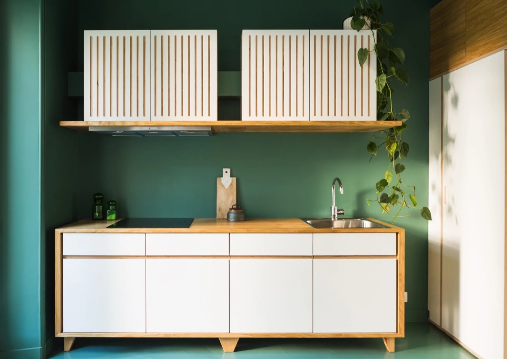 A modern minimalist kitchen features white upper cabinets with vertical slats, a warm wood countertop, and a dark green painted wall.