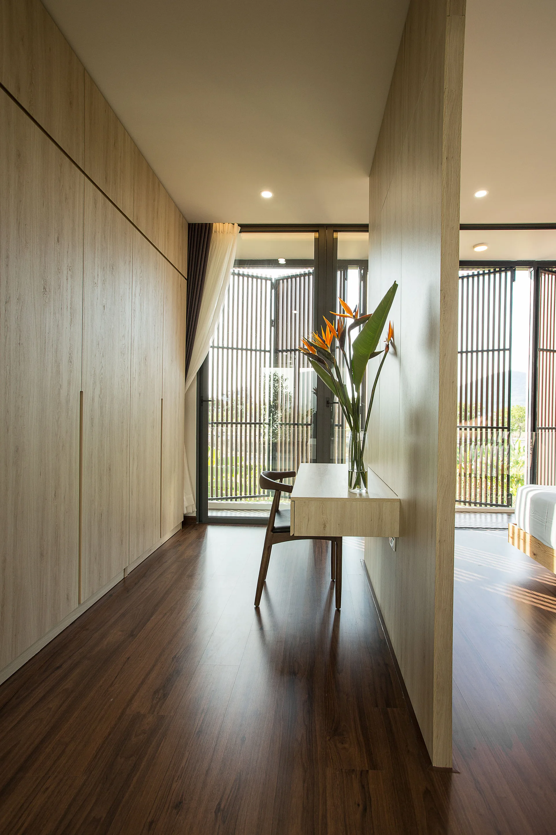 A floating wooden desk and dark wood chair sit against a partition wall next to a large light wood wardrobe in a modern minimalist room.