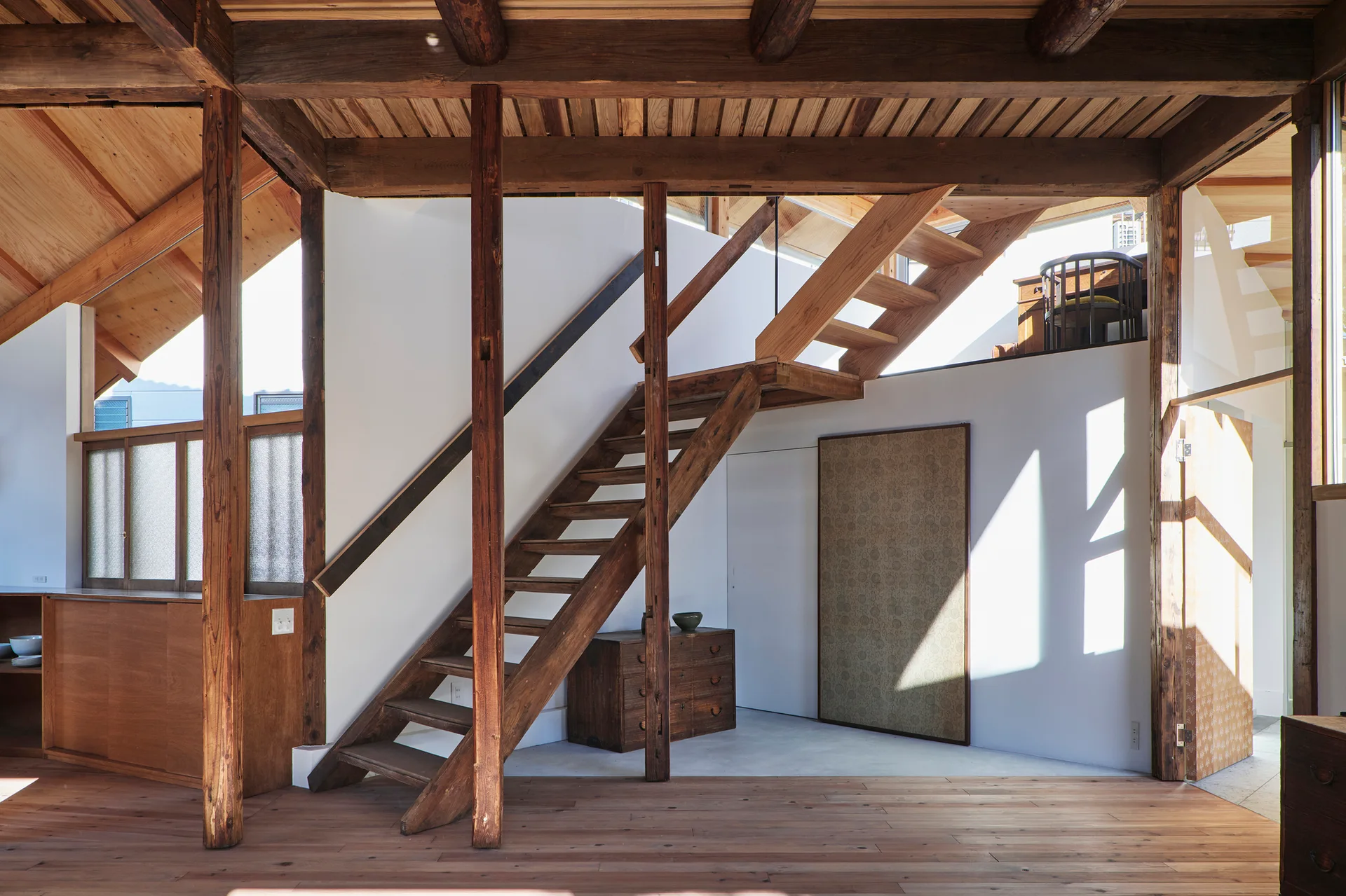 An open-tread wooden staircase ascends amid exposed timber posts and beams in a minimalist interior with white walls.