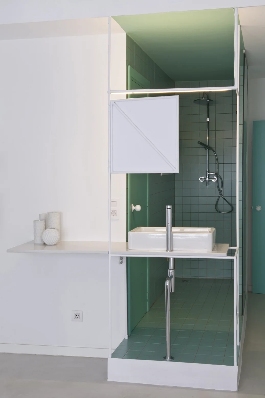 A minimalist bathroom features a white ceramic basin set on an open metal frame vanity, with green square tiled shower walls in the background.