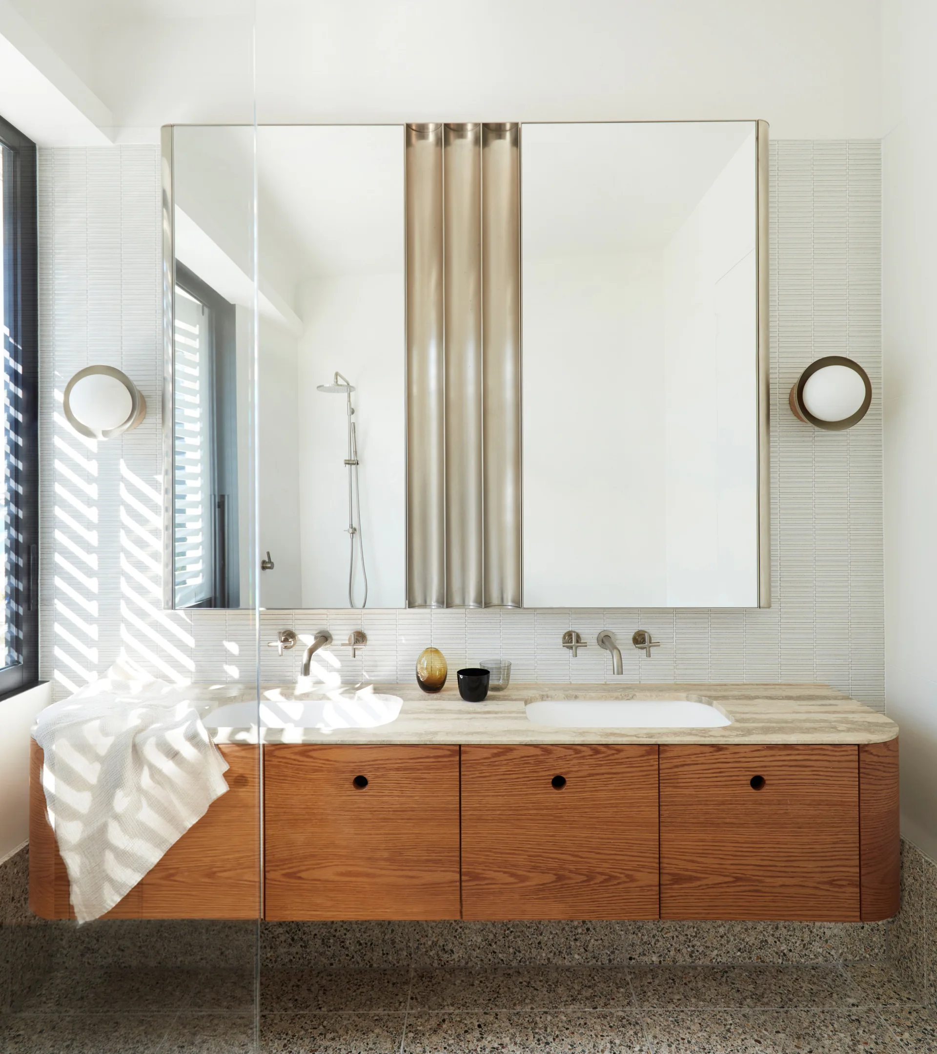 A modern bathroom features a floating wooden double vanity with beige stone countertops, integrated sinks, a mirrored cabinet, and stacked white wall tiles.