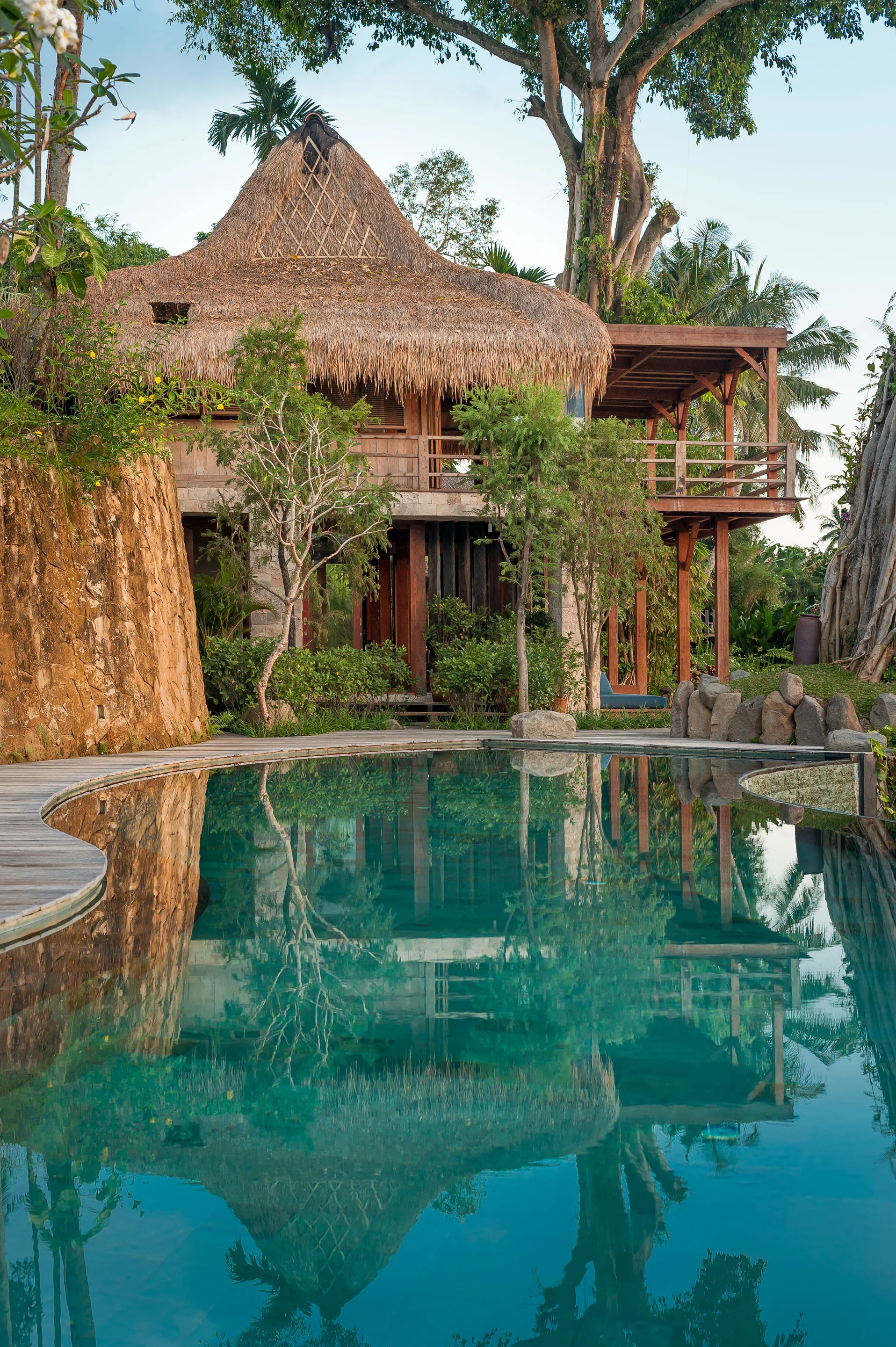 A two-story tropical wooden villa with a thatched roof stands beside a curved swimming pool with reflective green water and wooden decking.
