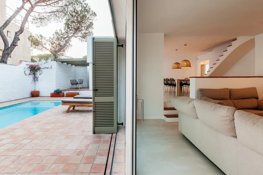 A modern indoor living room with a beige sofa opens up to a terracotta-tiled patio featuring a swimming pool and a green louvered shutter.