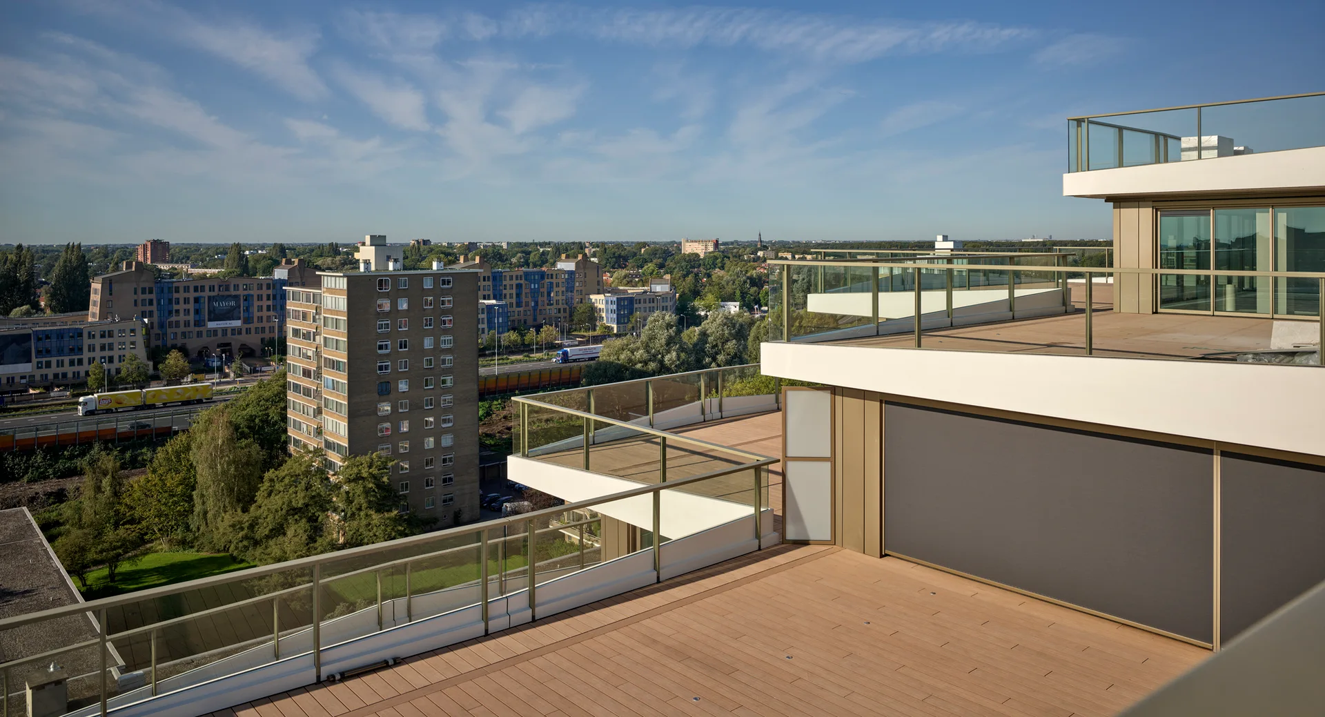 A modern multi-level apartment terrace features warm wood plank decking, minimalist glass railings, and retractable exterior shades.
