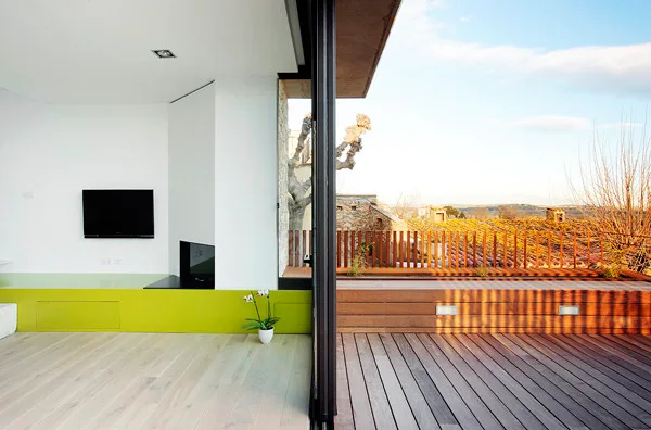 A modern indoor living area with light wood floors and a lime green low cabinet transitions through a glass sliding door to an outdoor wood plank deck.