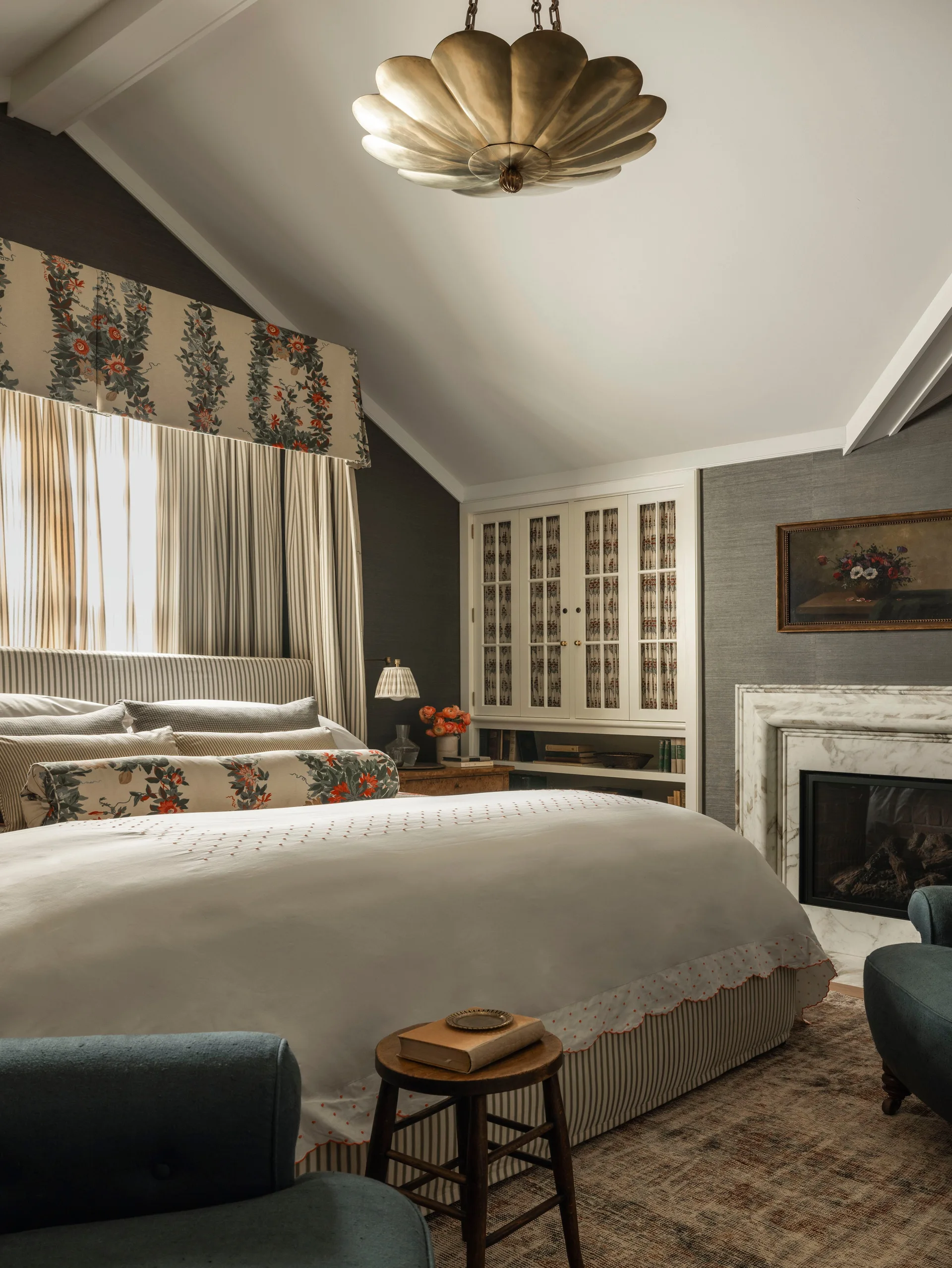 A traditional bedroom featuring a bed with striped and floral bedding, a scalloped brass ceiling fixture, and a white marble fireplace.