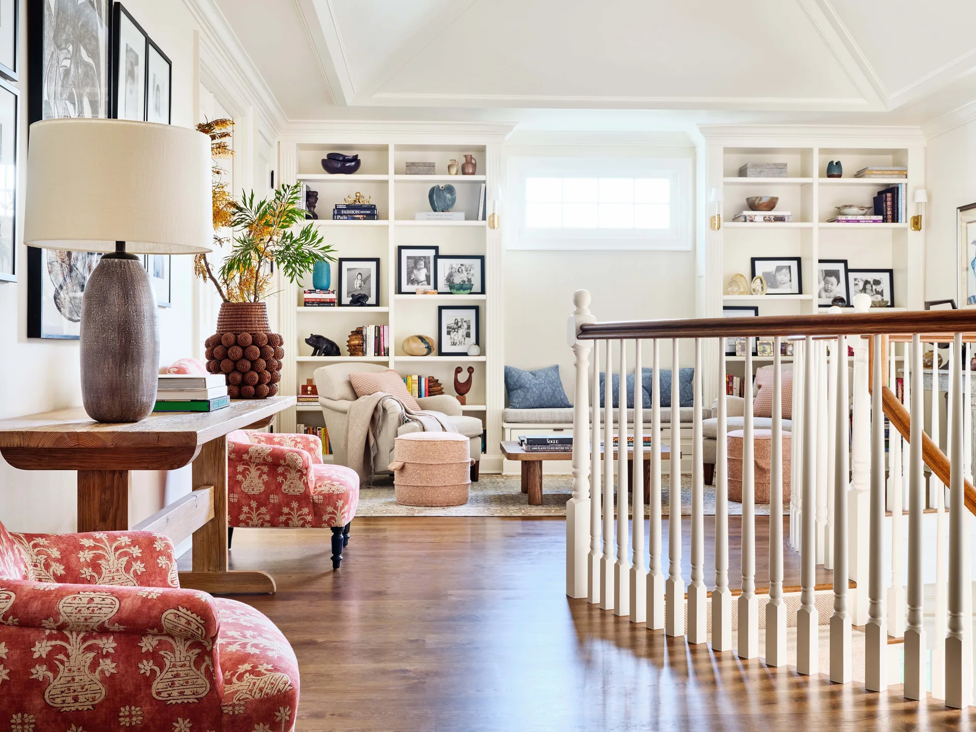 A wood-floored stair landing features a pair of red patterned armchairs, a wooden console table, and symmetrical built-in white bookshelves in the background.