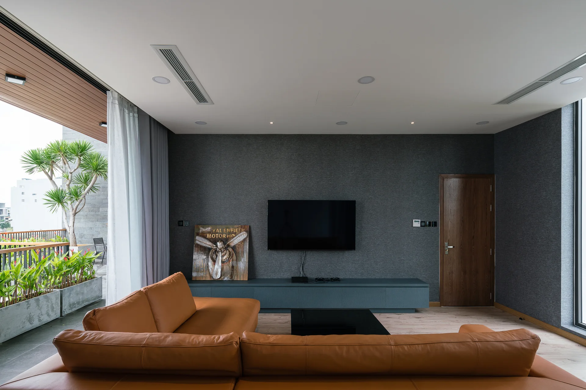 A tan leather sectional sofa faces a wall-mounted TV on a dark gray textured wall in a modern living room with balcony access.
