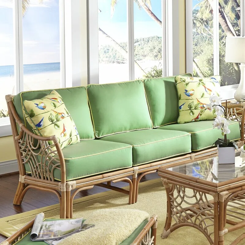 A green cushioned rattan sofa with bird-patterned pillows sits in a sunlit coastal room next to a matching rattan coffee table.