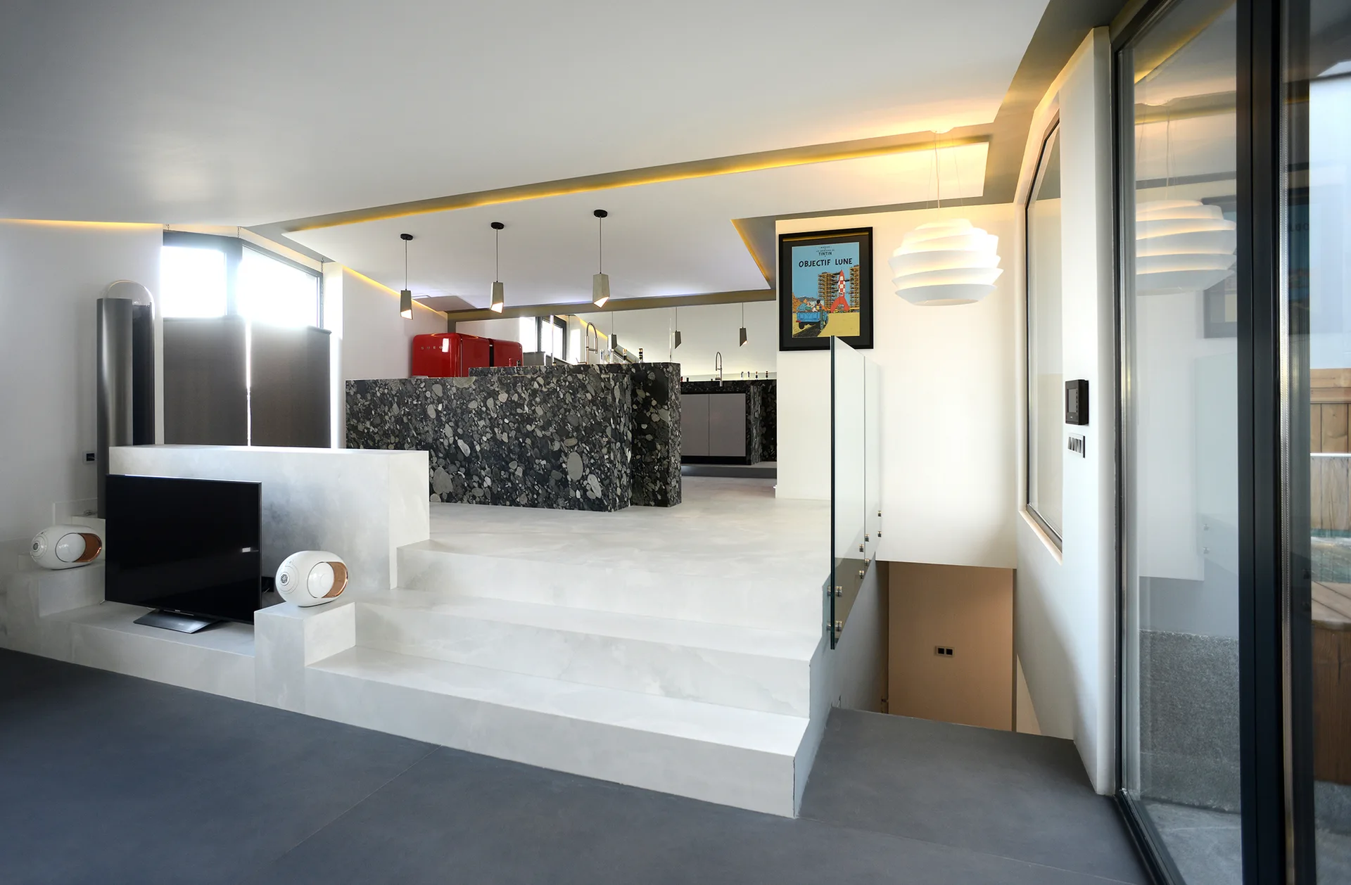 White tiered steps connect a lower media room to an upper level featuring a dark speckled stone kitchen island and a red refrigerator.