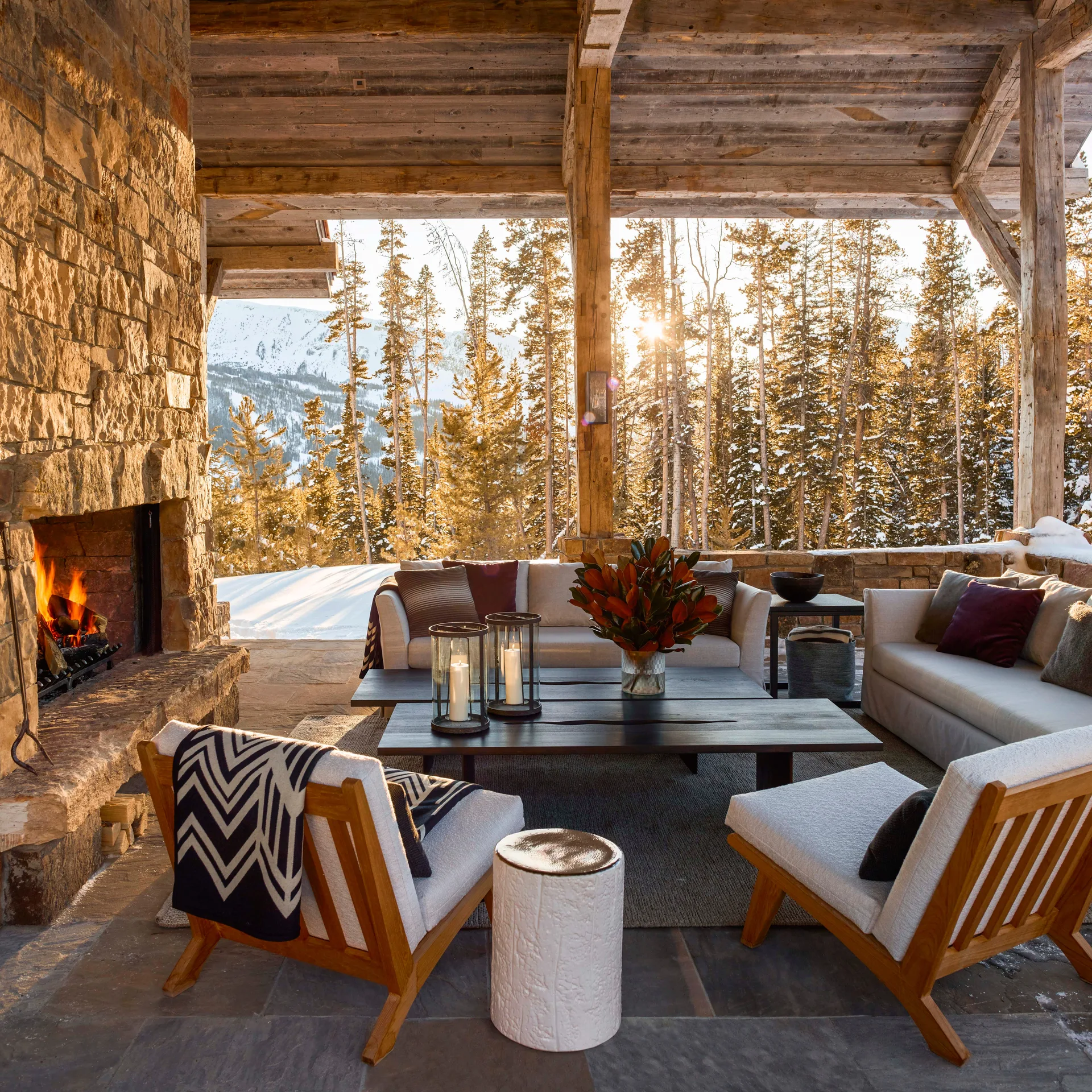 A rustic covered patio features a stone fireplace, timber beams, off-white sofas, and wood lounge chairs overlooking a snowy pine forest.