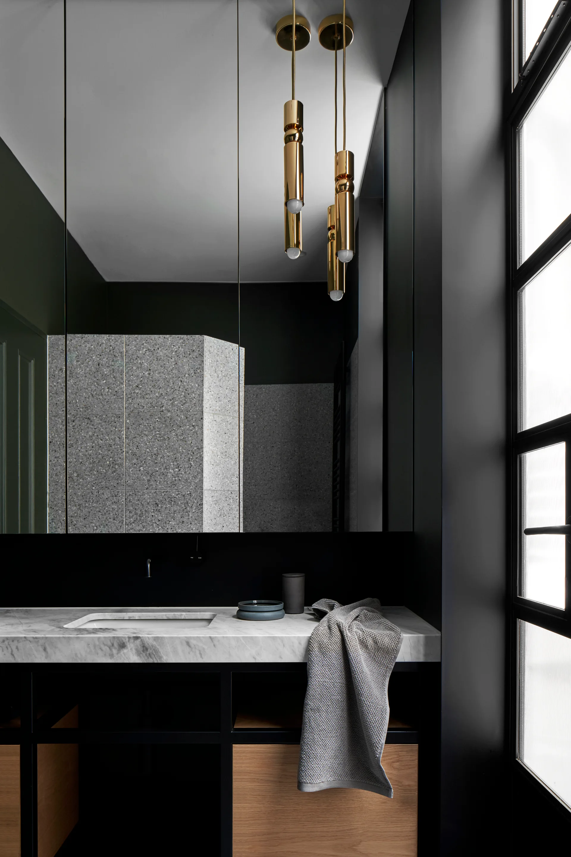 A modern bathroom vanity features a white marble countertop, light wood drawers under a black frame, and gold cylinder pendant lights.