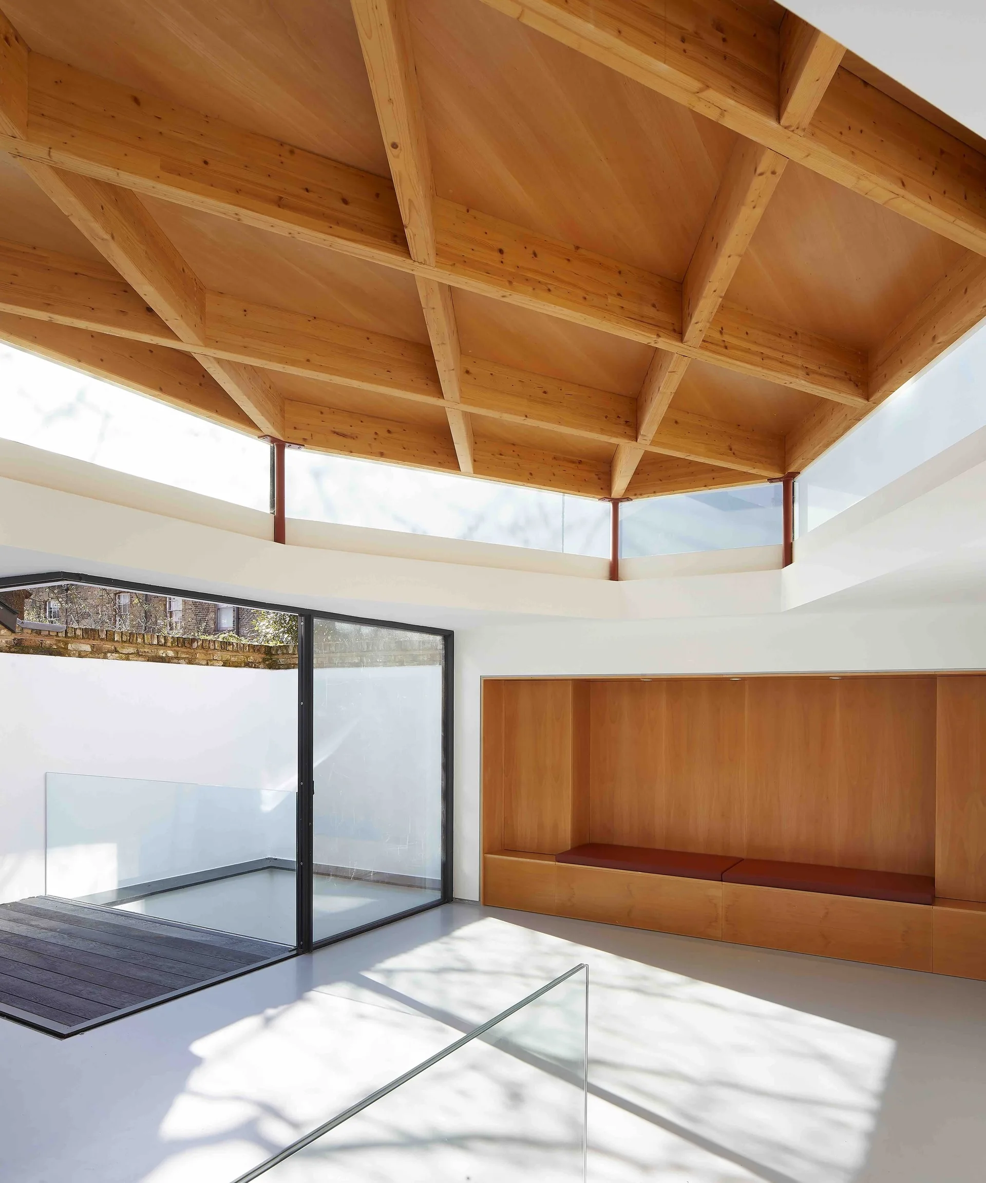 A minimalist room featuring a geometric timber grid ceiling, a built-in wooden bench alcove, and large glass sliding doors.