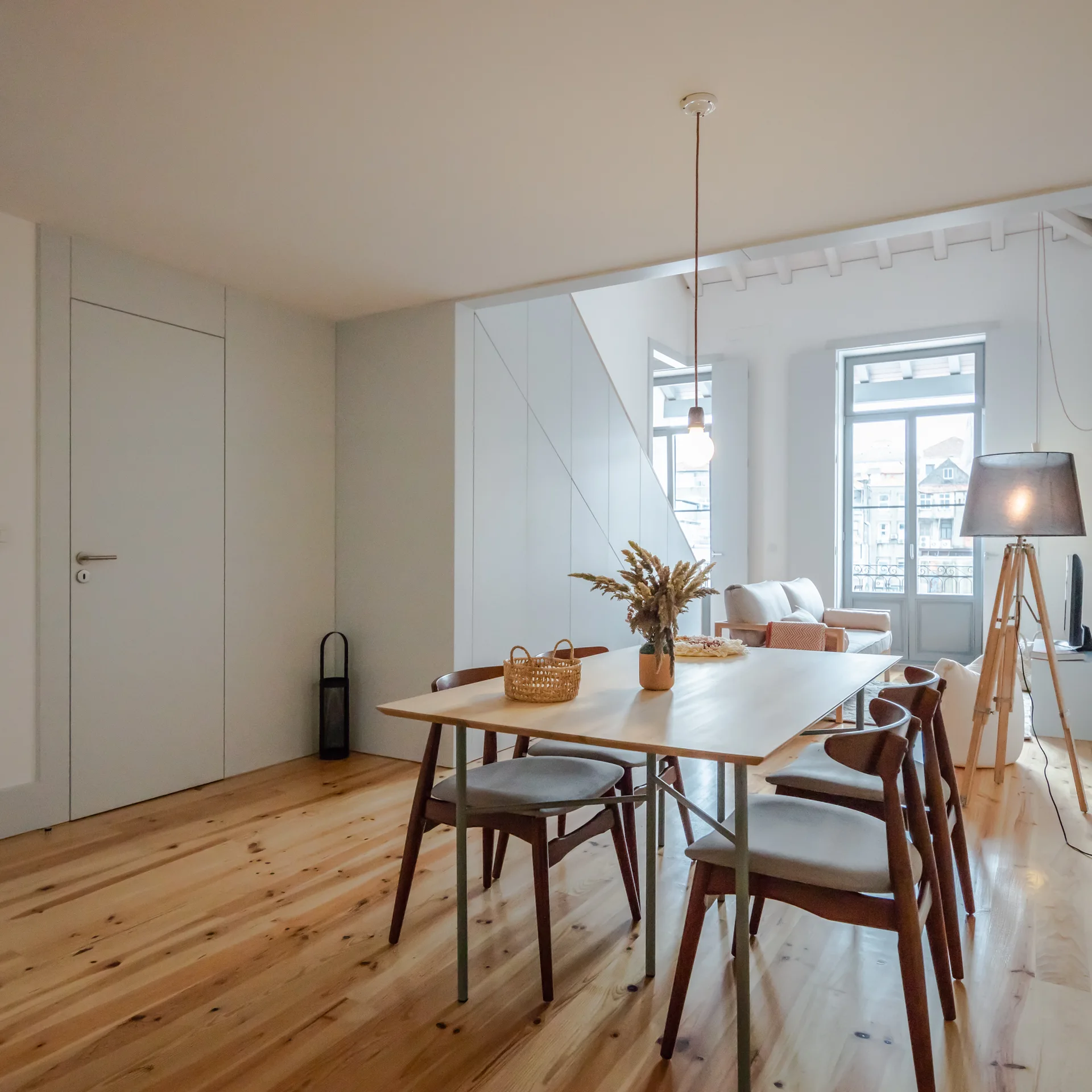 A minimalist dining room featuring a light wood table, grey upholstered chairs, natural pine floors, and an integrated white flush door.