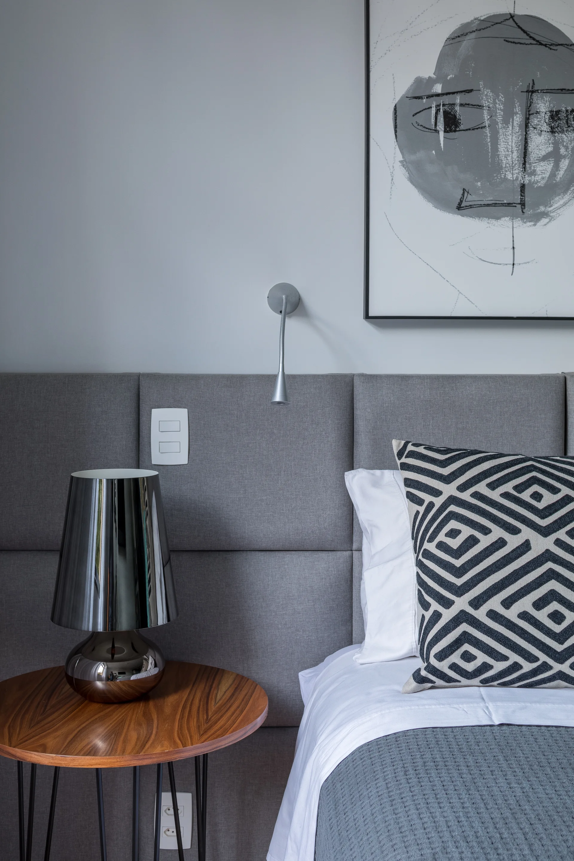 A gray upholstered headboard stands behind a round wood side table with a reflective chrome lamp and a bed styled with a geometric pillow.