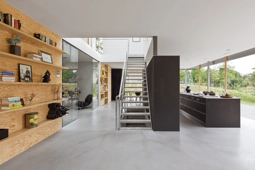 A minimalist open-plan interior featuring a custom plywood shelving wall, a central metal staircase, and a dark kitchen island over concrete floors.