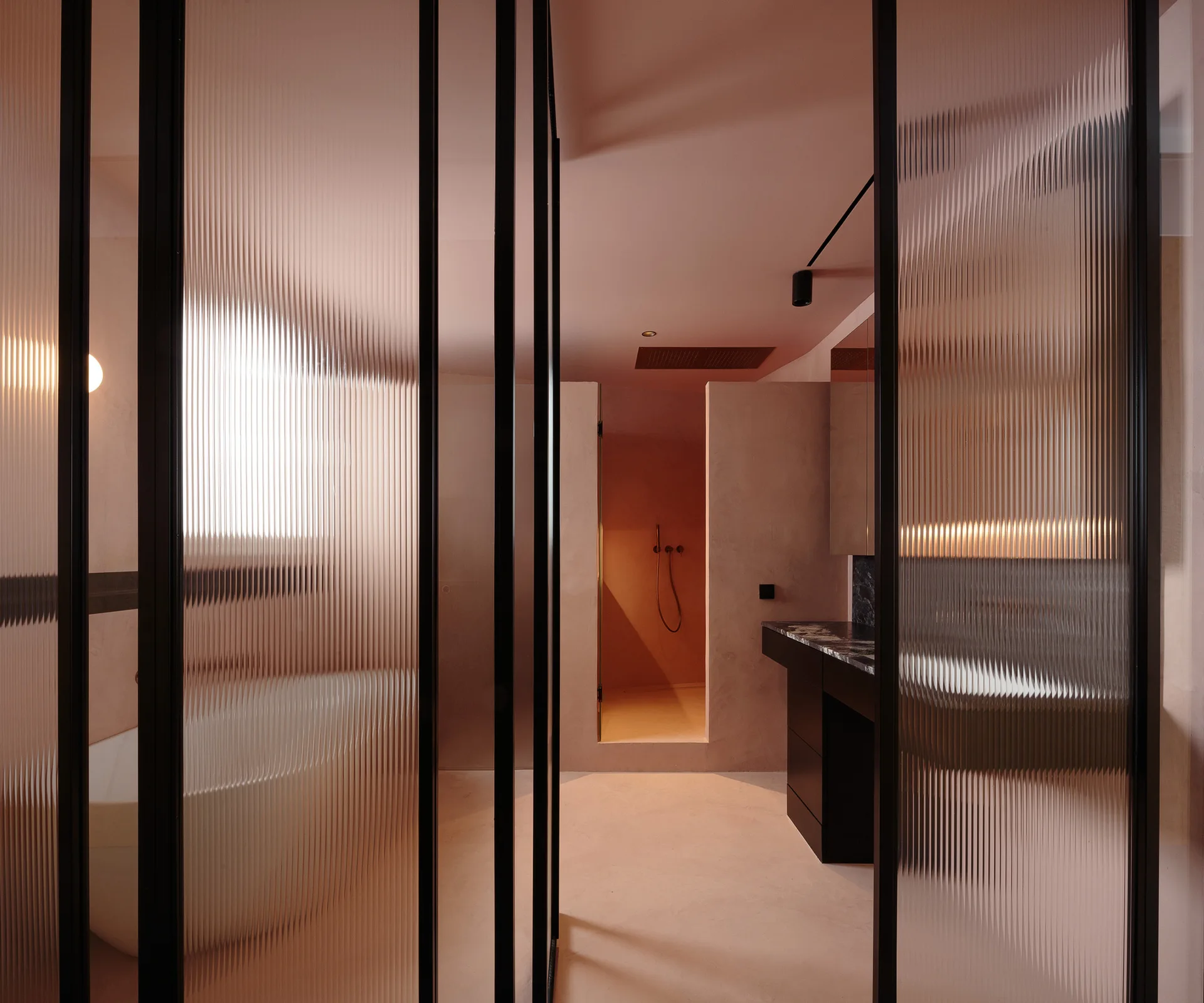 A minimalist pink bathroom features fluted glass room dividers in black frames, a white freestanding tub, and a dark marble vanity.