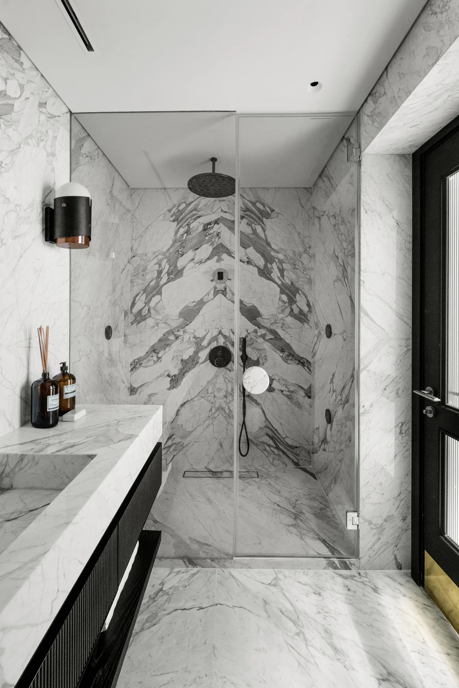A modern bathroom features a glass shower stall with book-matched veined marble walls, an integrated marble vanity sink, and a black framed door.
