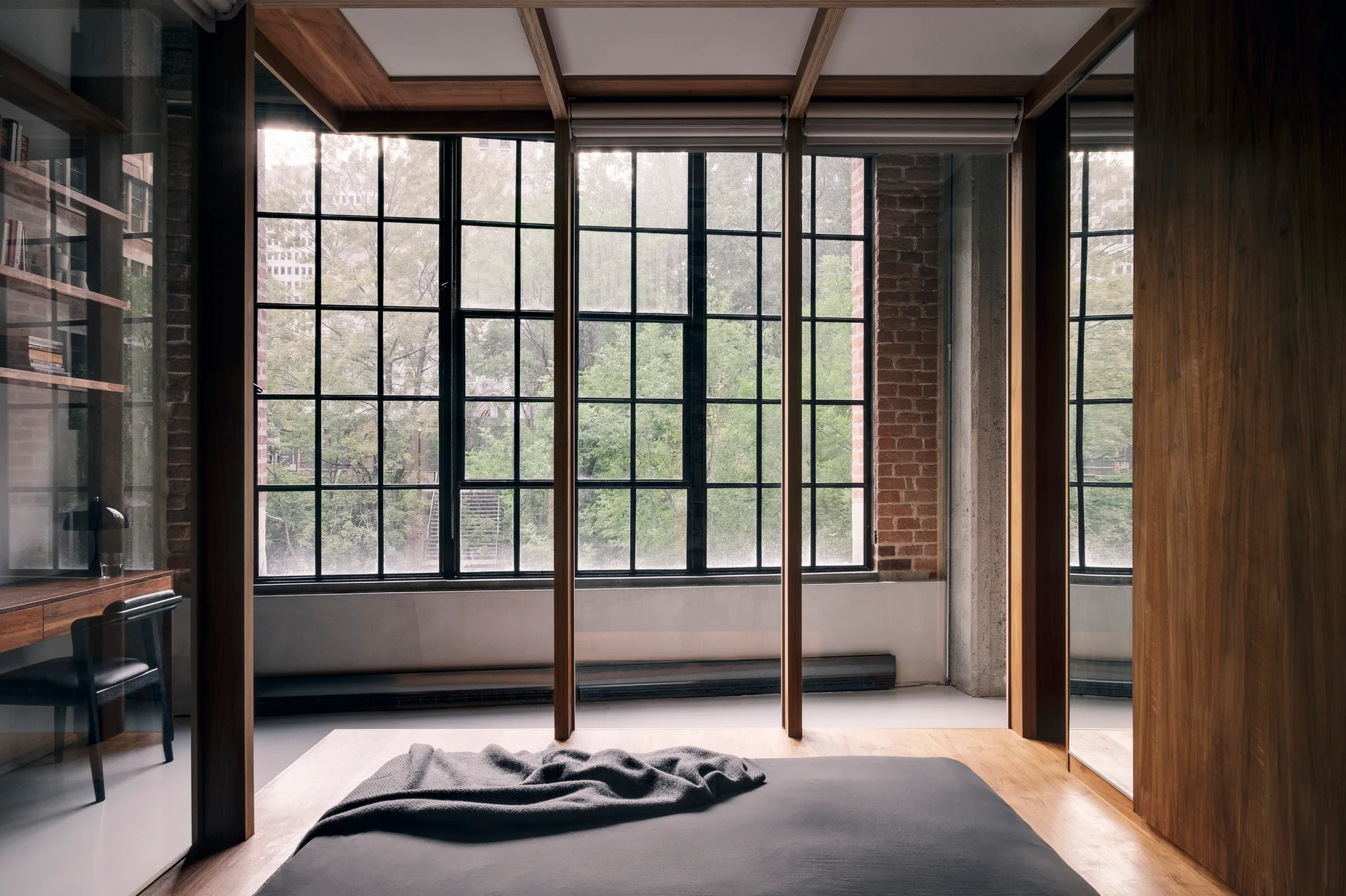 A modern platform bed faces a large black grid window in a bedroom featuring exposed brick and a glass-enclosed study space.