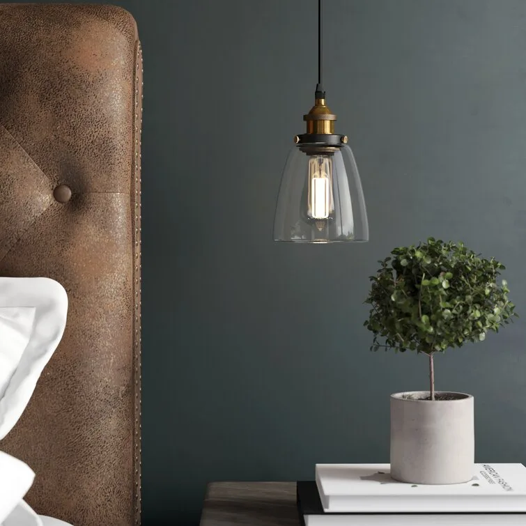 A glass pendant light with a warm filament bulb hangs beside a tufted brown leather headboard and a small potted plant on a nightstand.