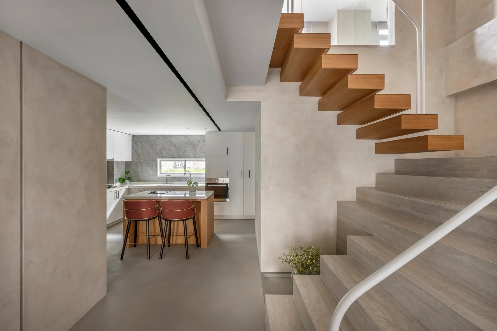 A modern floating wood staircase stands next to textured plaster walls, with a kitchen island and two red bar stools in the background.