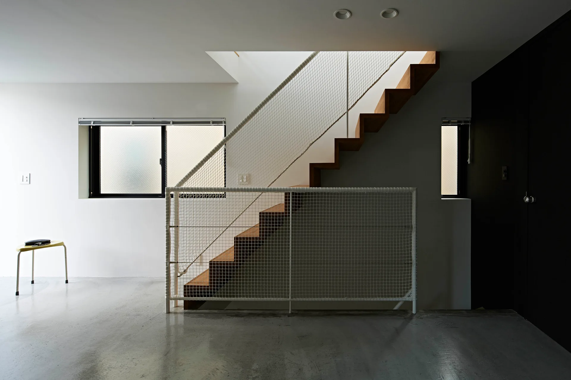 A wooden floating staircase with a white net safety railing stands on a polished concrete floor against plain white walls.