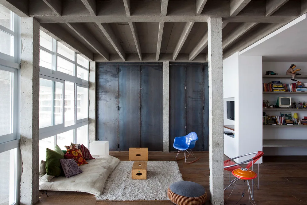 A modern loft living room features an exposed concrete grid ceiling, a low floor cushion sofa, wooden floors, and dark steel wall panels.