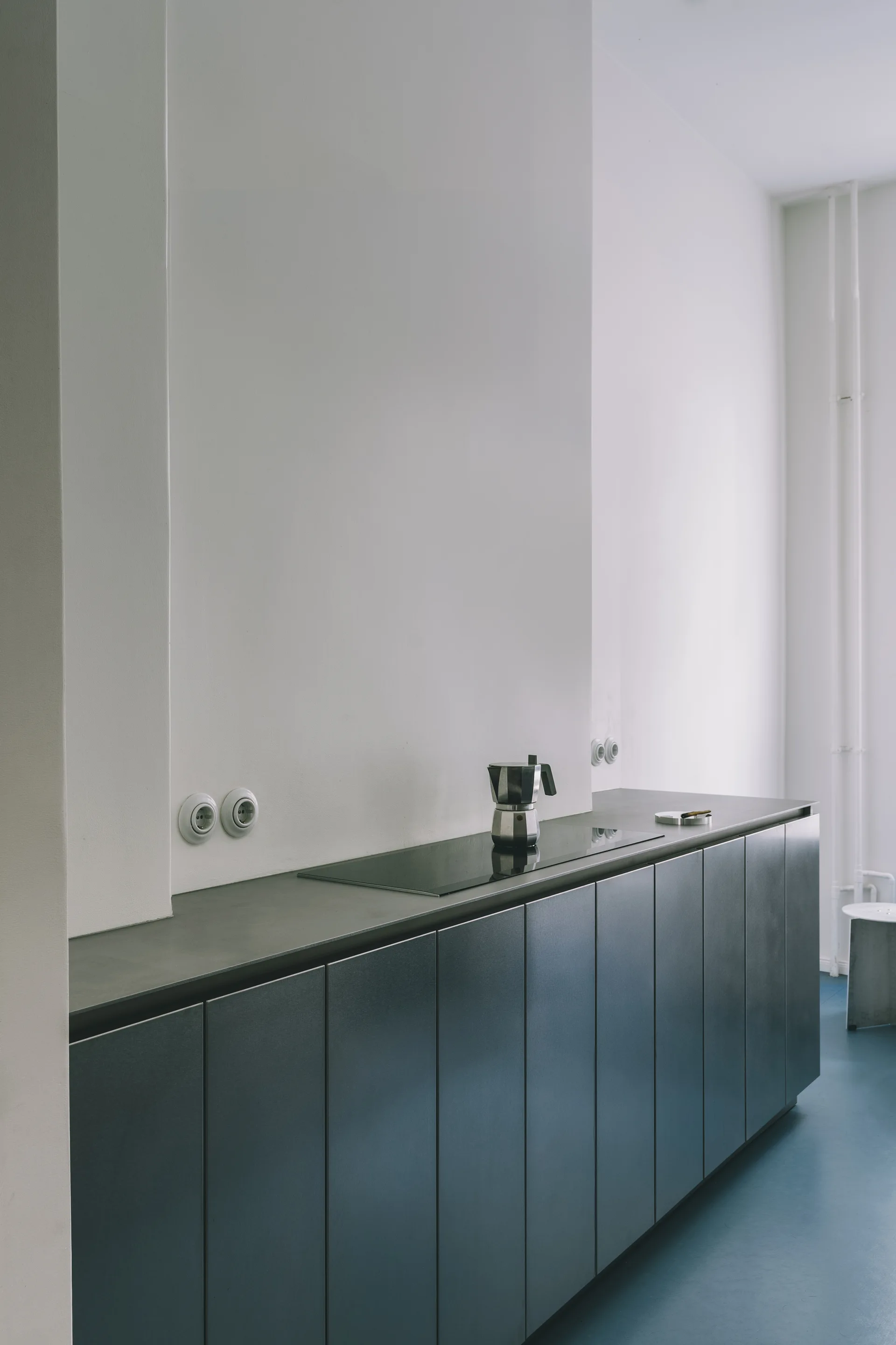 A minimalist kitchen features sleek dark gray metal cabinets, a concrete countertop with a glass cooktop, and a smooth blue floor against white walls.