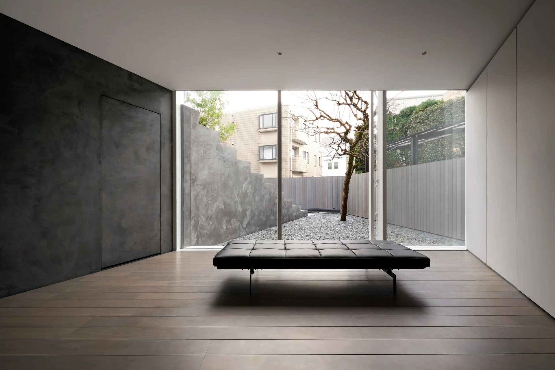 A minimalist room features a black tufted leather daybed positioned before a floor-to-ceiling glass window looking out onto a gravel courtyard.