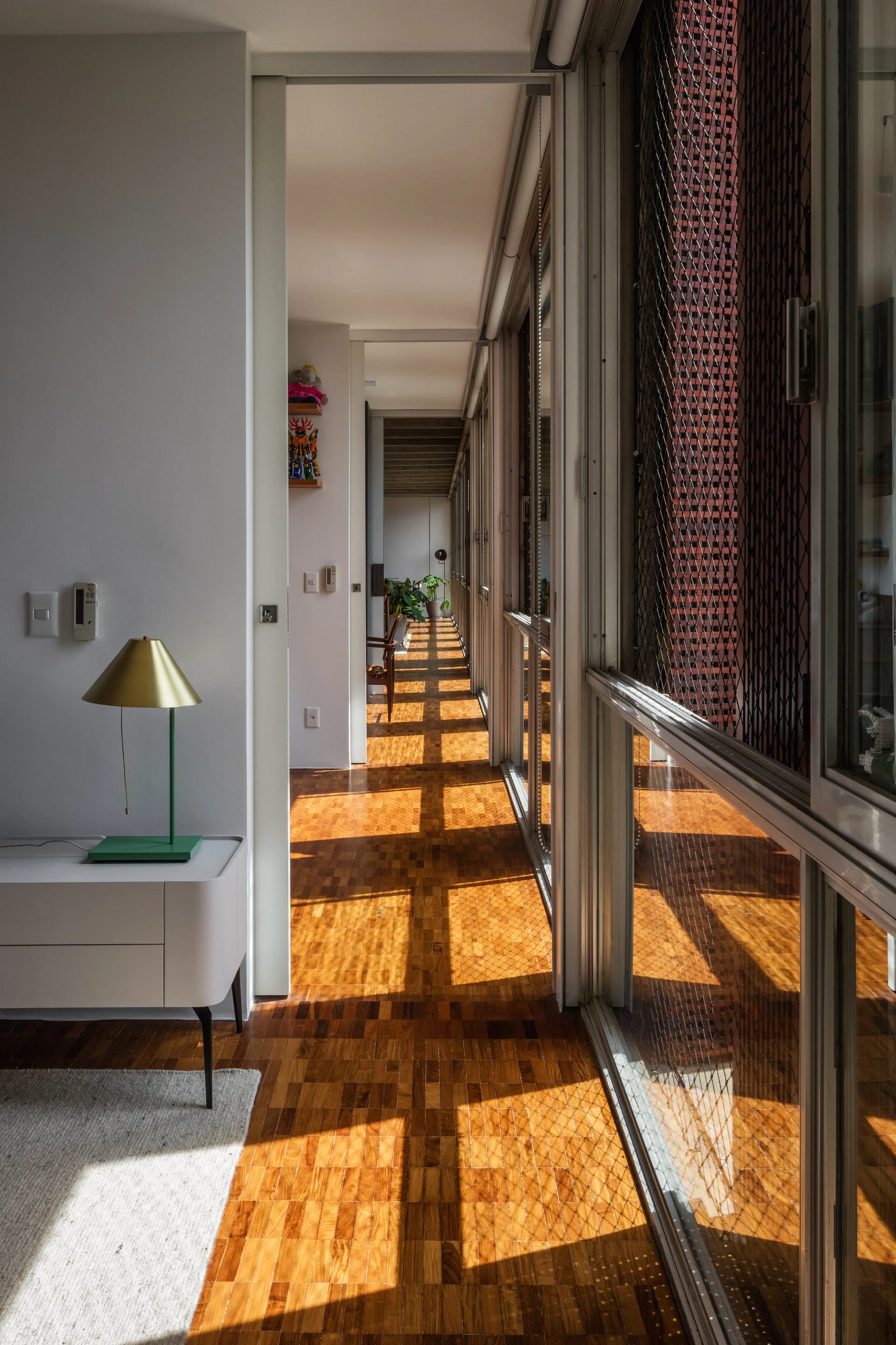 A long sunlit hallway features polished wooden parquet flooring, sliding glass doors, and a green table lamp resting on a white console table.