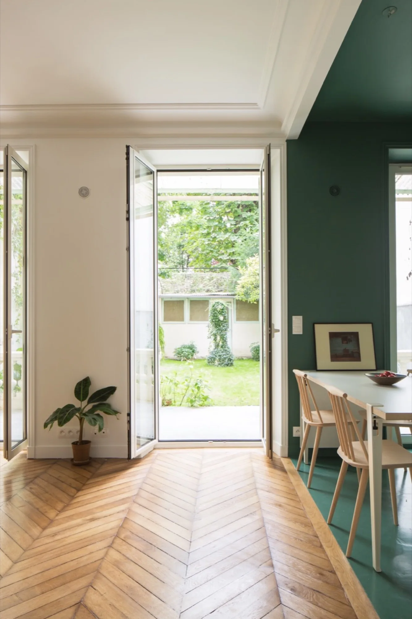 A dining room featuring herringbone wood floors, a green accent wall, and wood chairs next to open French doors leading to a green backyard.