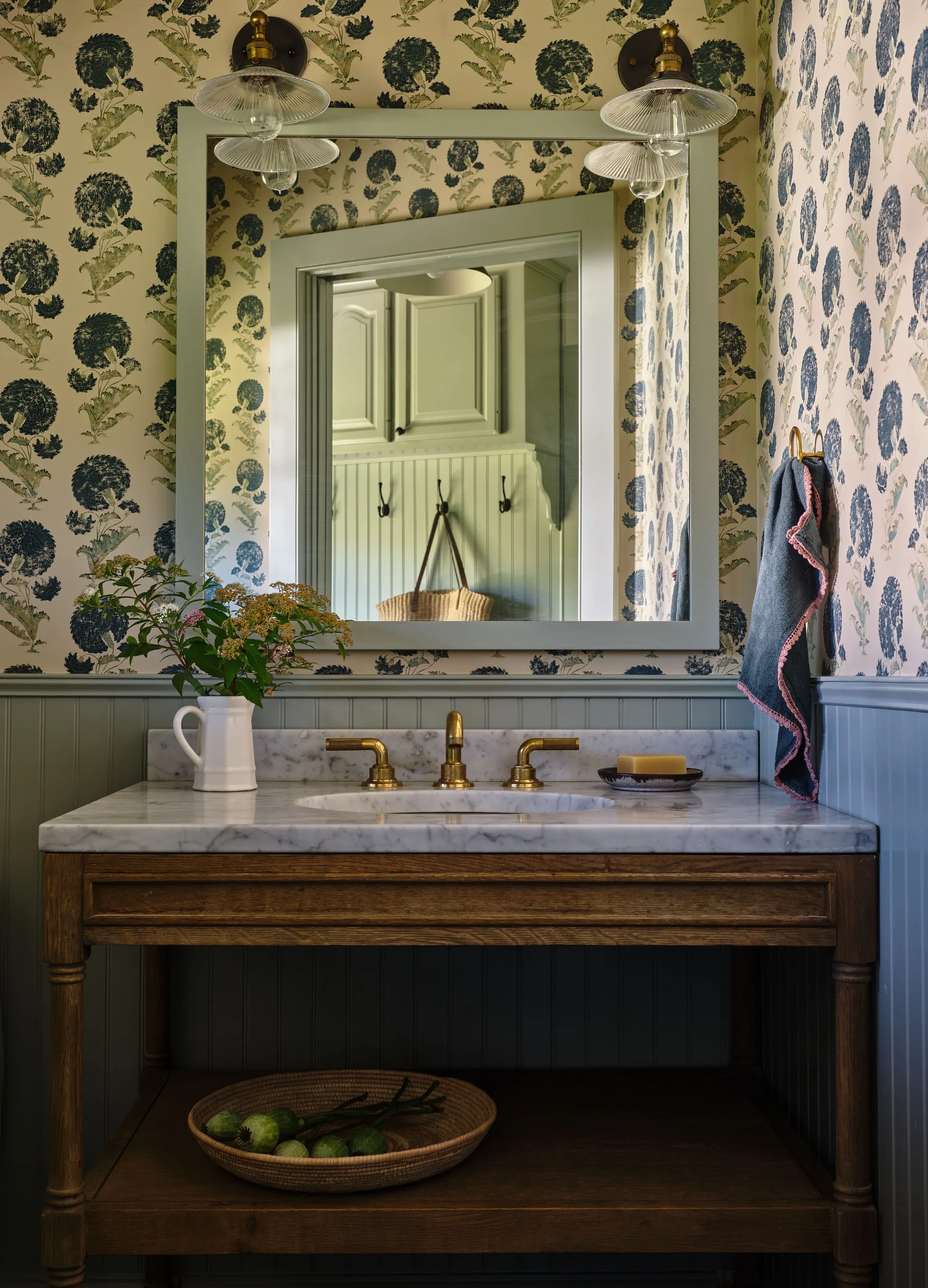 A wood bathroom washstand with a marble top stands against sage green beadboard wainscoting and botanical wallpaper.