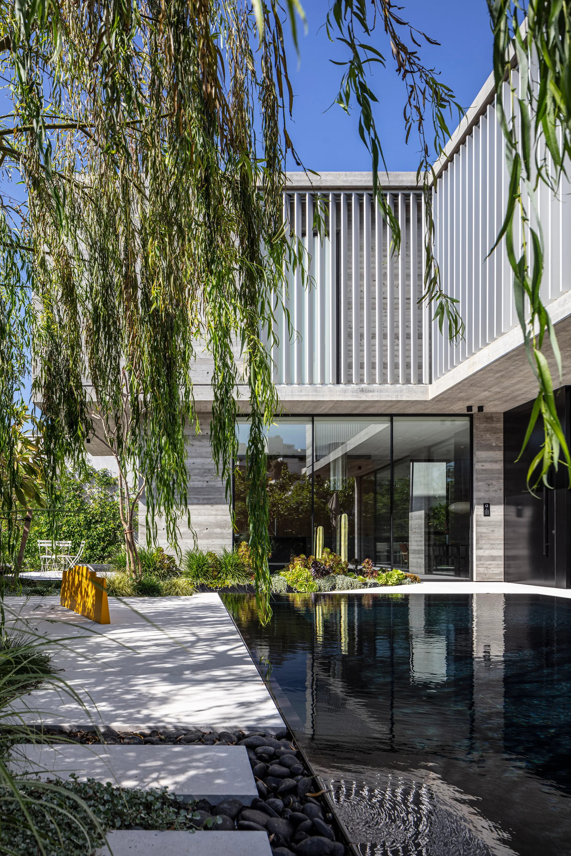 A modernist concrete home overlooks a dark swimming pool in a courtyard shaded by a weeping willow and featuring a bright yellow bench.