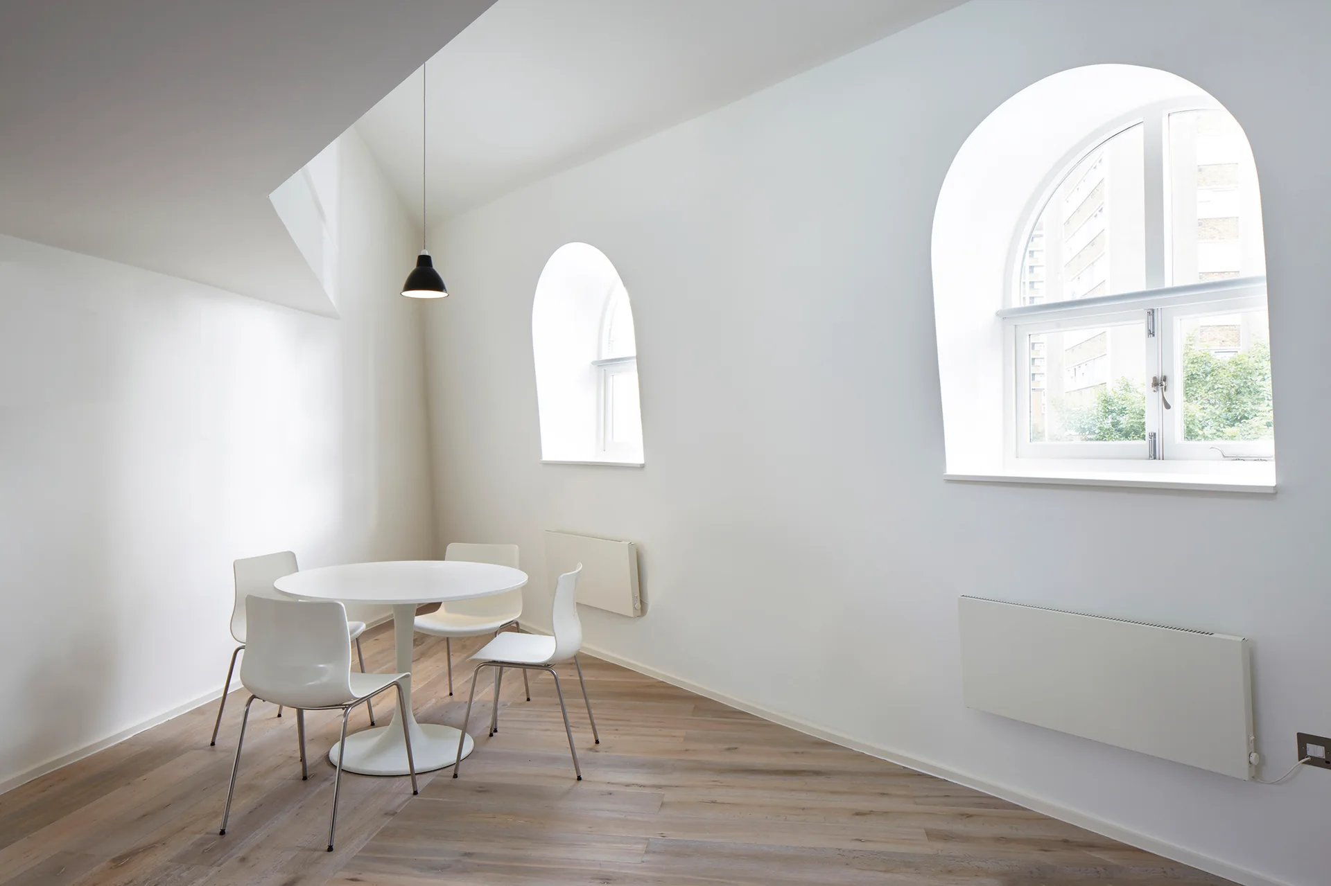 A minimalist white dining room features a round pedestal table, white chairs, and two deep arched windows overlooking trees.