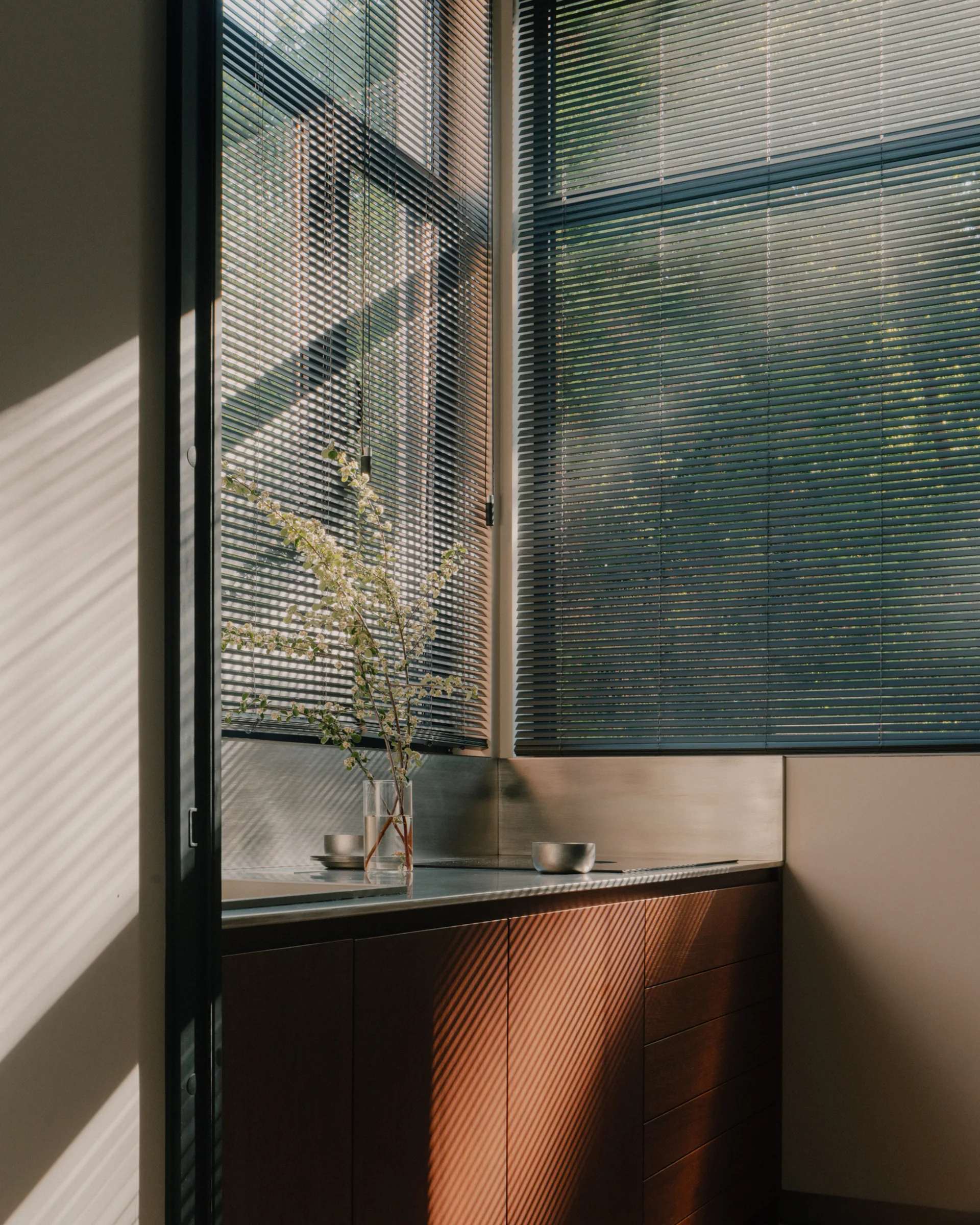 A corner kitchen features warm wood cabinetry, a metallic backsplash, and venetian blinds casting linear shadows.
