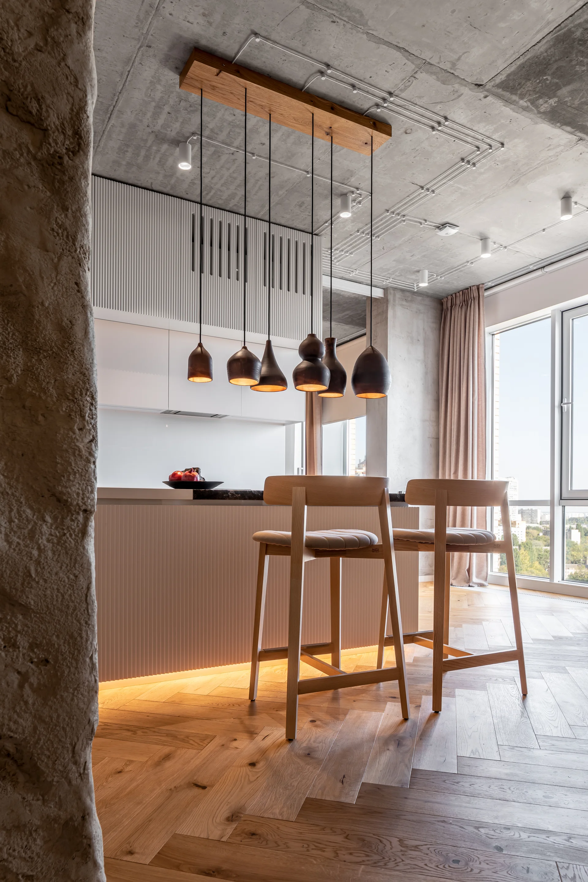 A modern kitchen features a white ribbed island with wooden bar stools, a row of dark pendant lights, and an exposed concrete ceiling.