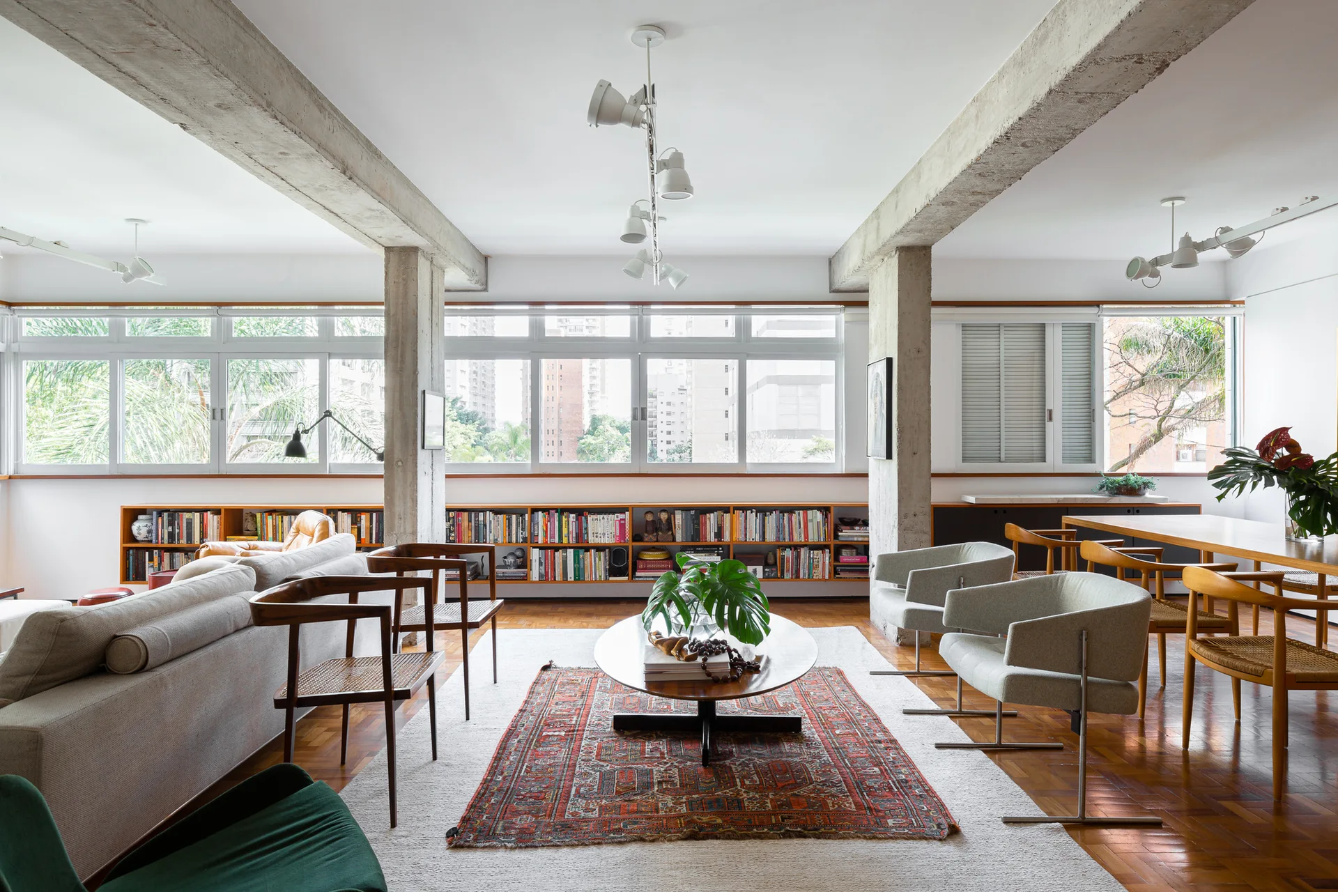 A bright open-plan living room featuring exposed concrete beams, a long low-profile bookshelf beneath wide windows, and mid-century modern seating.