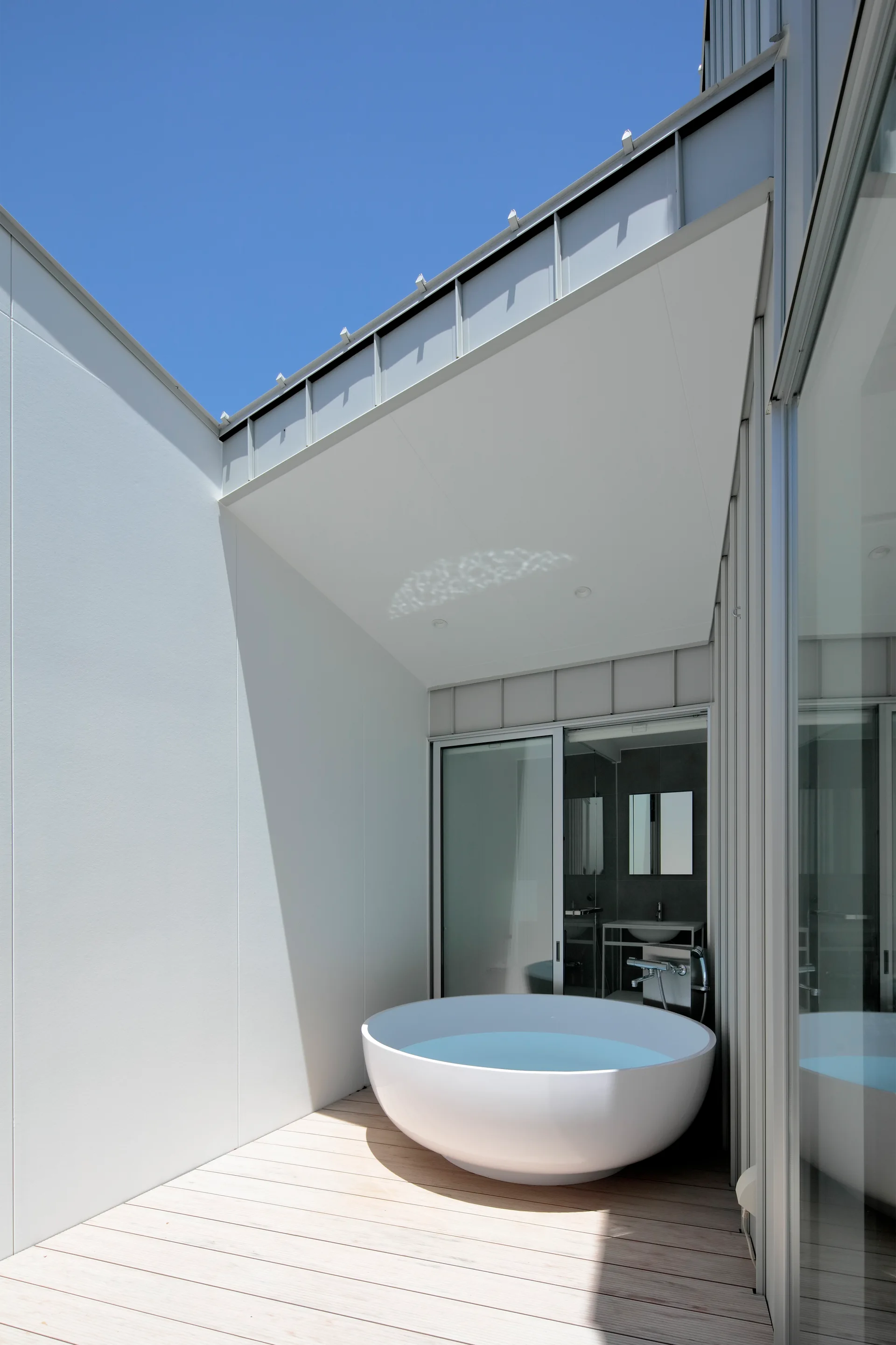 A circular white freestanding bathtub sits on a light wood deck on a modern outdoor patio next to a glass sliding door.