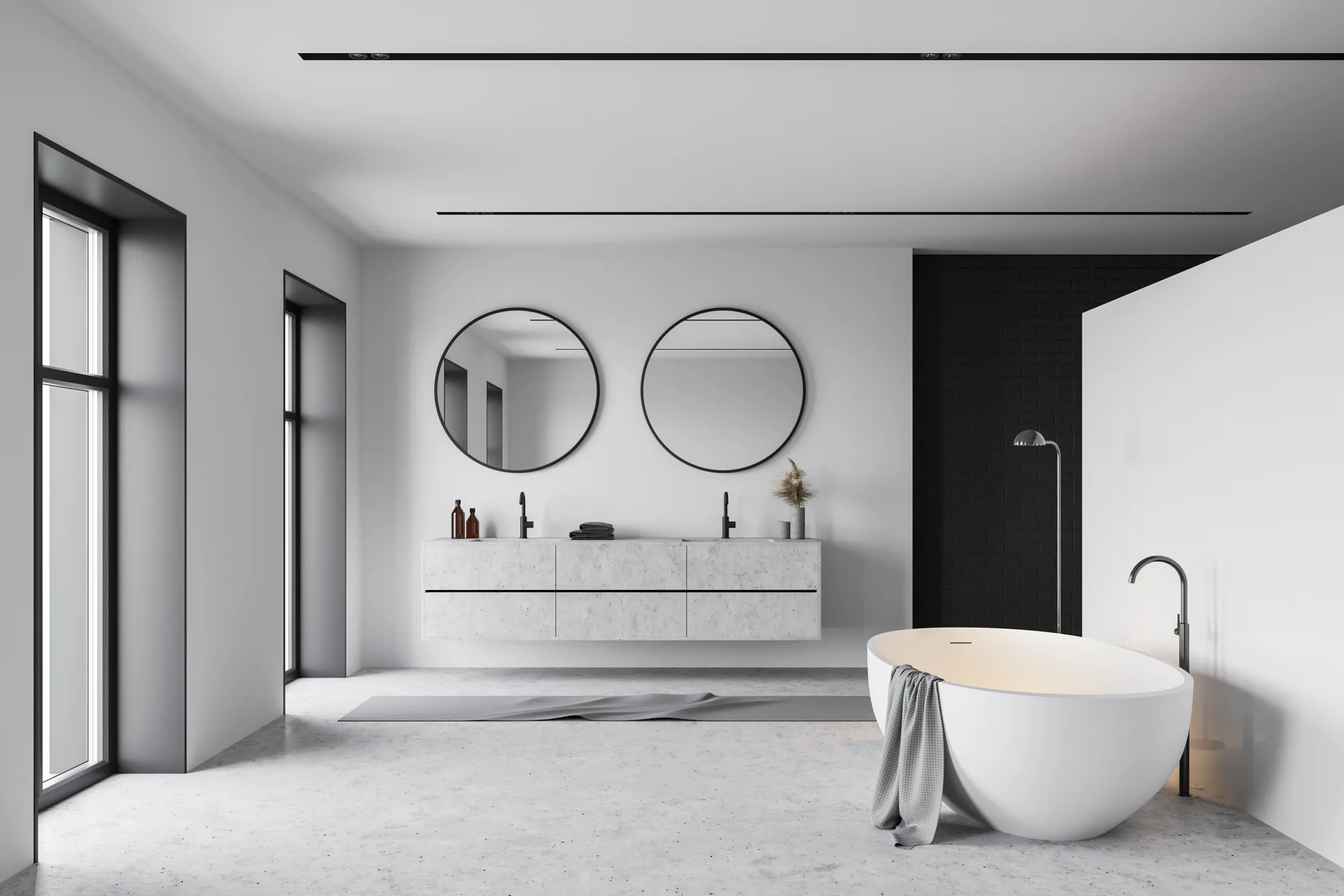 A minimalist bathroom features a floating stone vanity with twin round mirrors, a freestanding white bathtub, and a black brick accent wall.