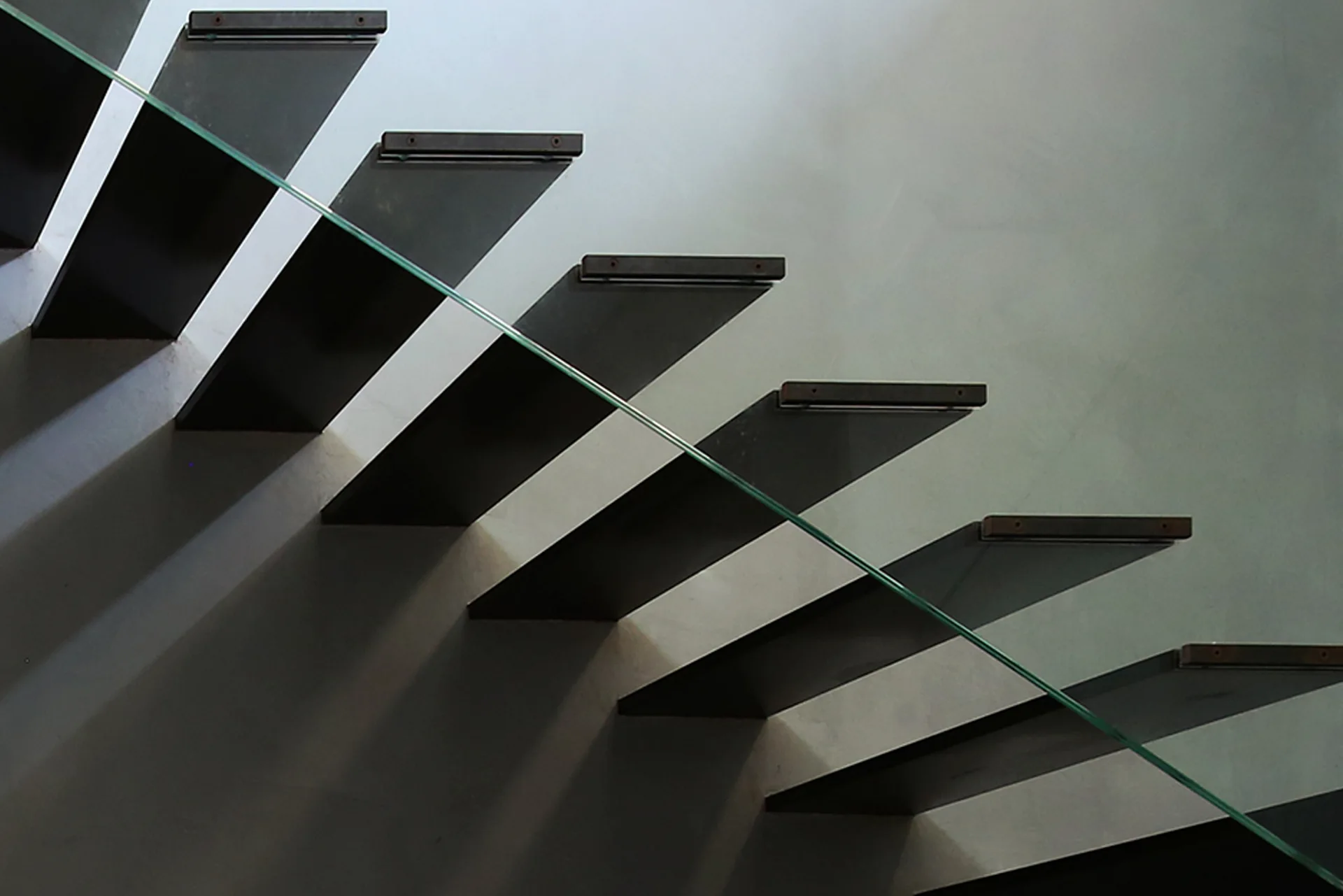 A close-up of dark minimalist floating stair treads casting angled geometric shadows onto a light grey wall next to a glass railing panel.