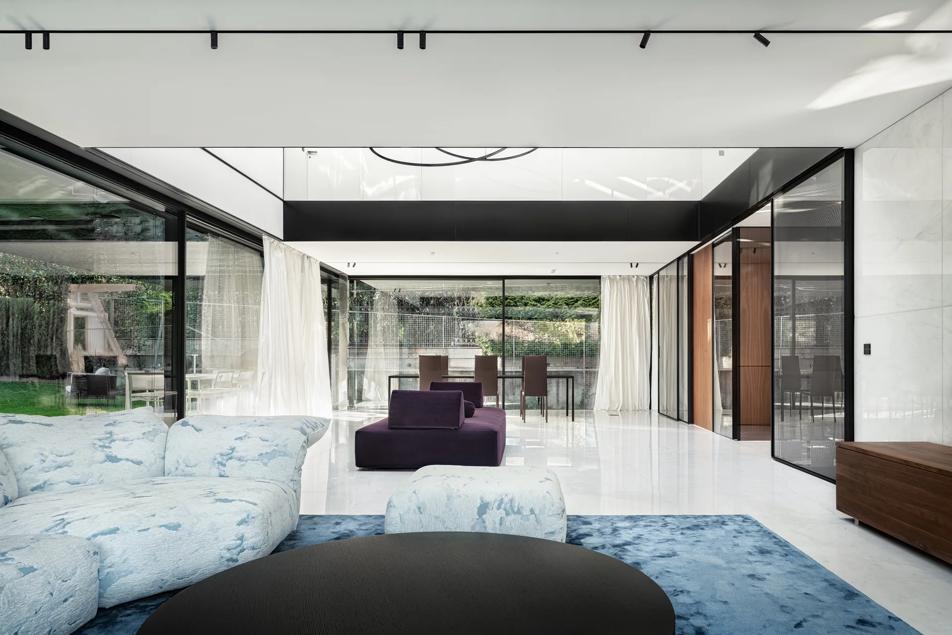 A minimalist living room with a patterned blue sofa, a purple velvet-like sofa, and glass walls opening to an outdoor yard.