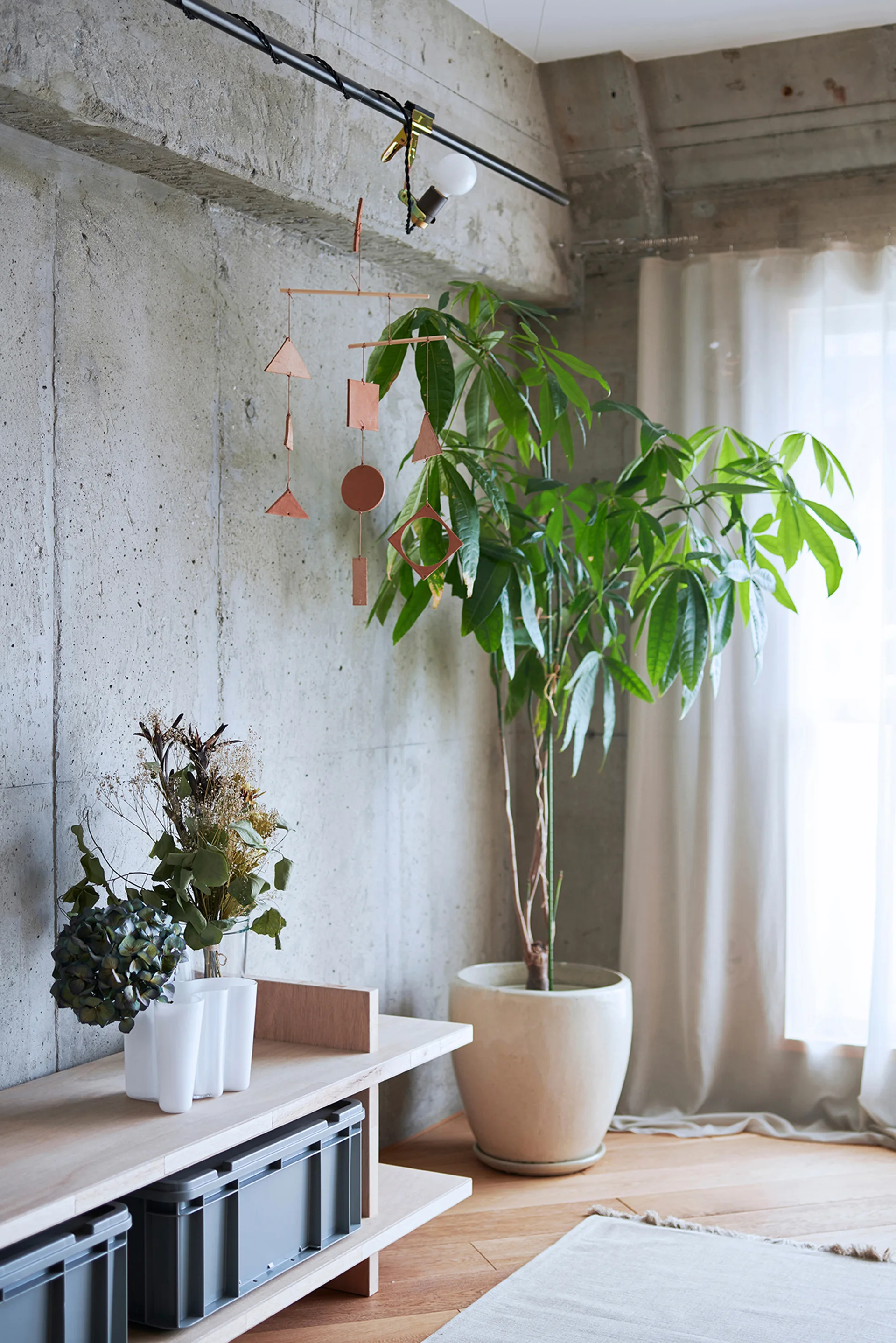 A light wood console table sits against an exposed concrete wall next to a large potted plant and a geometric hanging mobile in a bright room.
