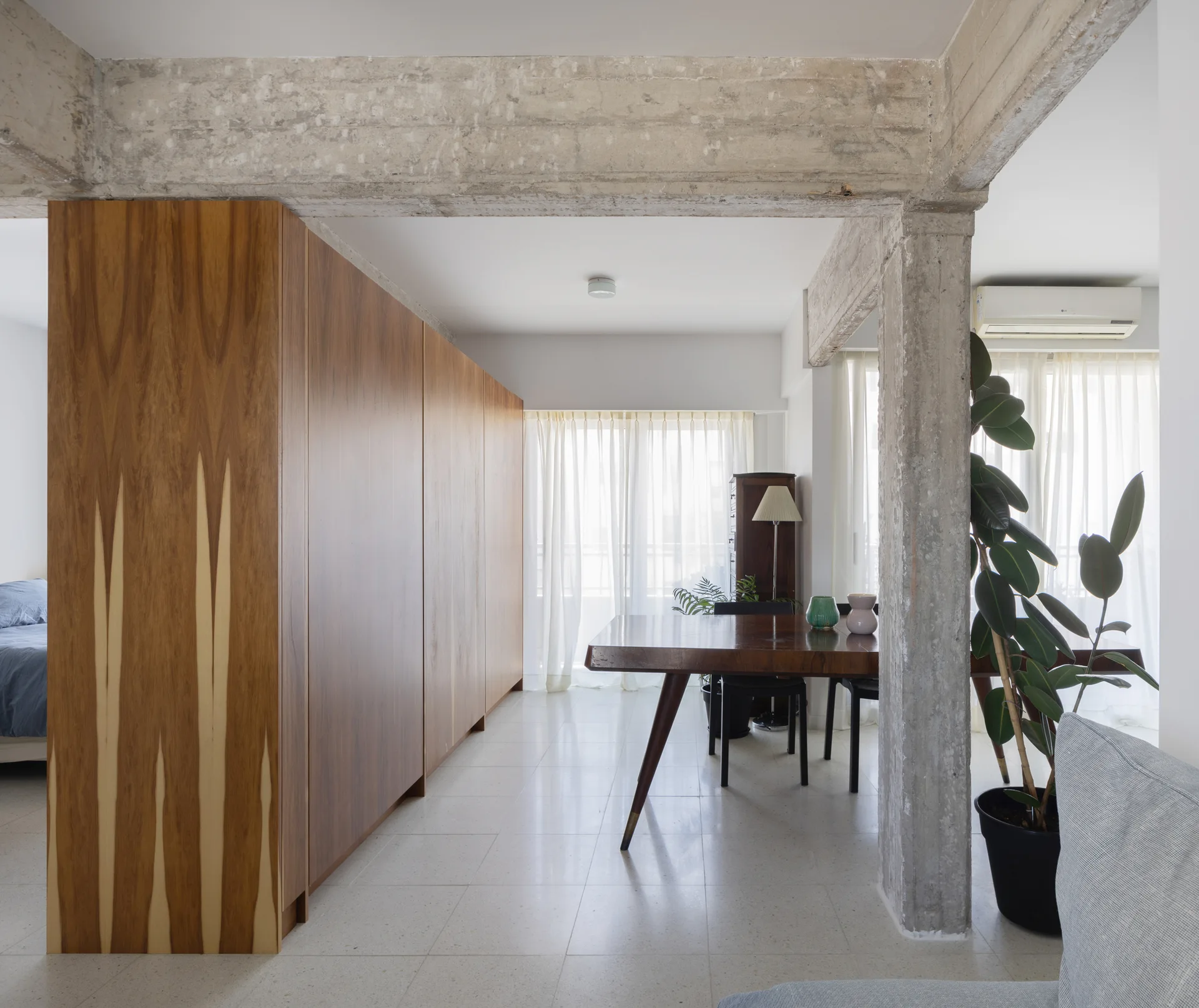 A modern dining room features an exposed concrete pillar and beam next to a large wood cabinet and a mid-century wooden table with chairs.